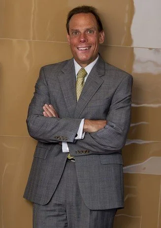 Jonathan Sablone standing with arms crossed, smiling in a gray suit with a yellow tie, against a beige wall.
