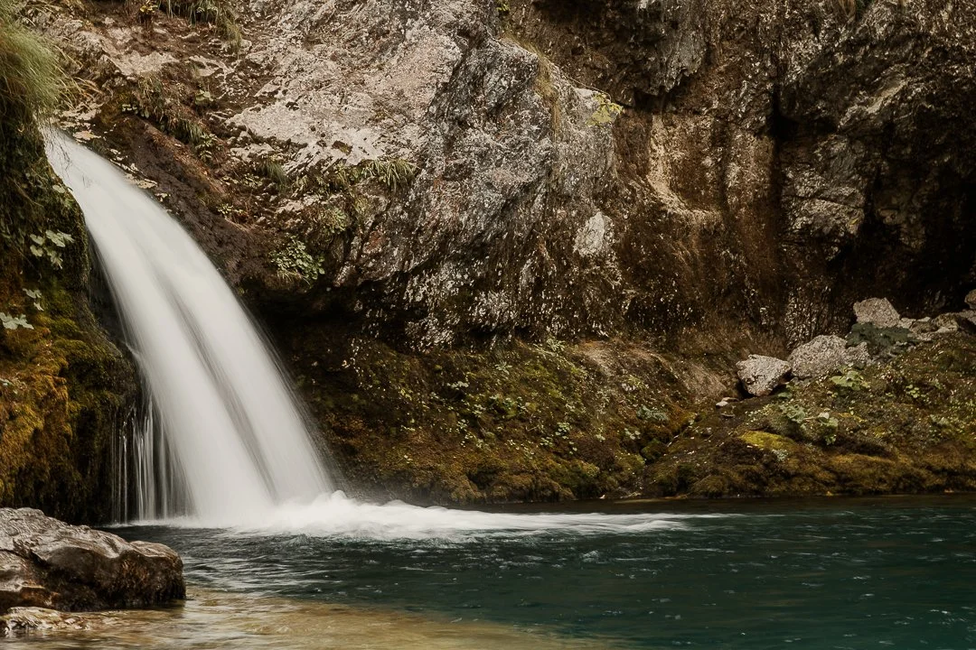 a slow exposure white waterfall shoots into a turquoise blue pool of water surrounded by mossy white limestone rocks in the albanian alps near Theth, the blue eye of theth
