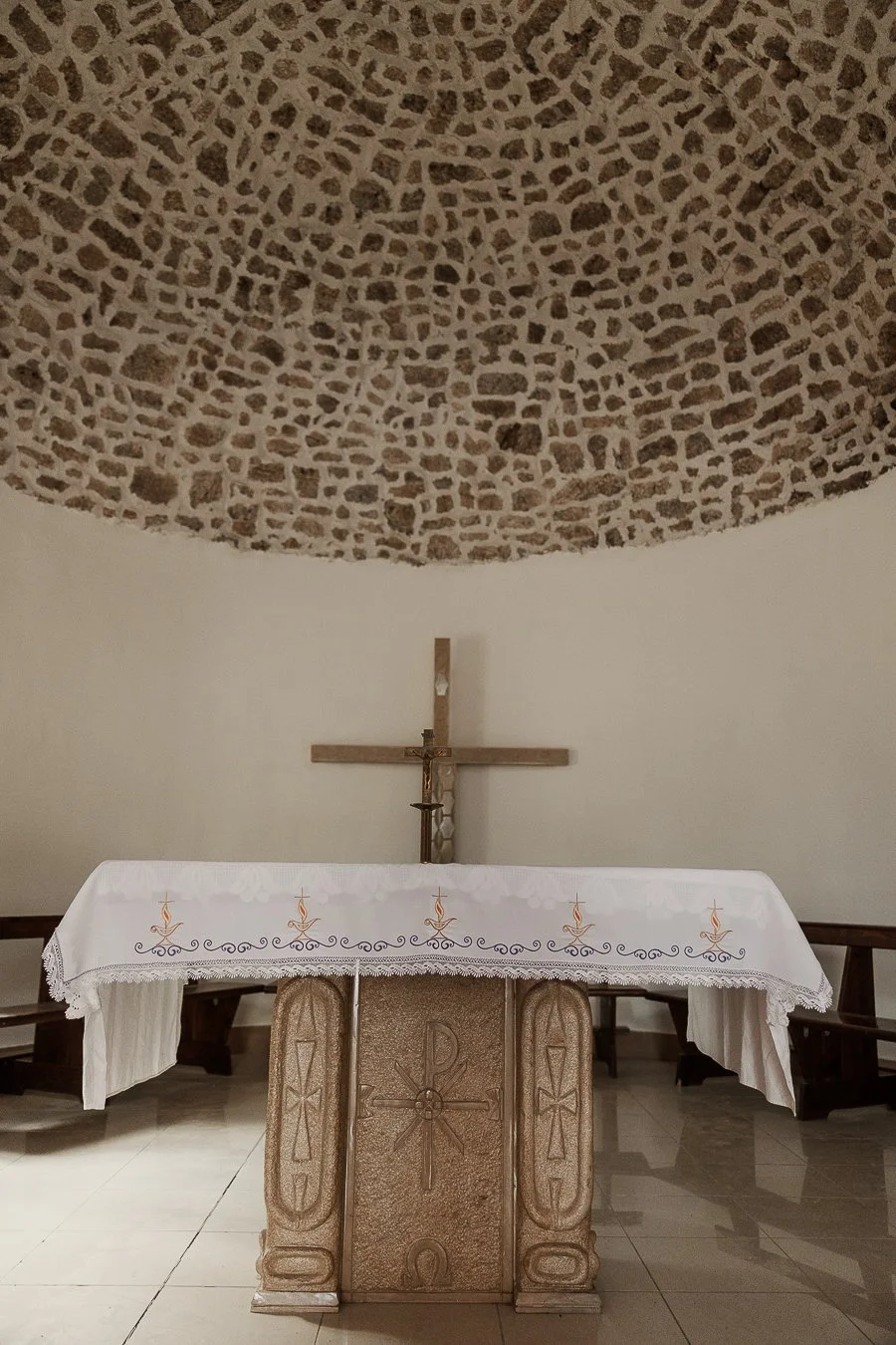 A carved pale stone altar under a rough stone domed ceiling in the small chapel of Theth, Albania, with a white embroidered cloth and a wooden cross.
