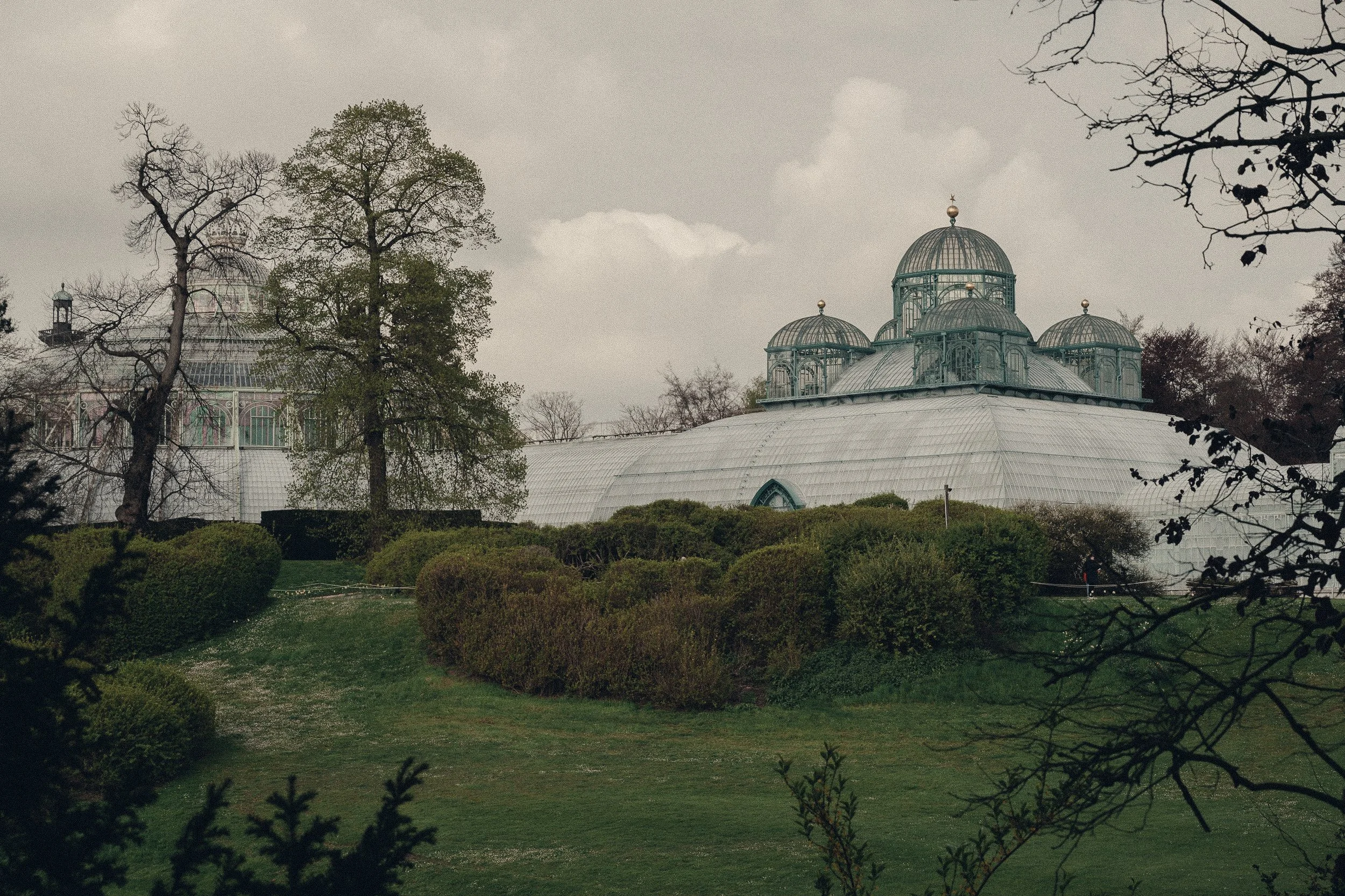 A large glass greenhouse with multiple domes, surrounded by trees and bushes, under a cloudy sky.