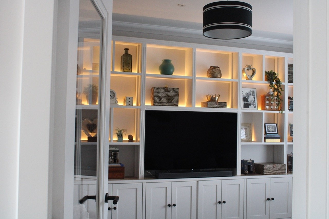 White built-in shelving in a handmade, traditional media wall with a large flat-screen TV in the centre. The shelves are illuminated with warm backlighting and integrated LEDs with soundbar integrated. Built-in cupboards. 