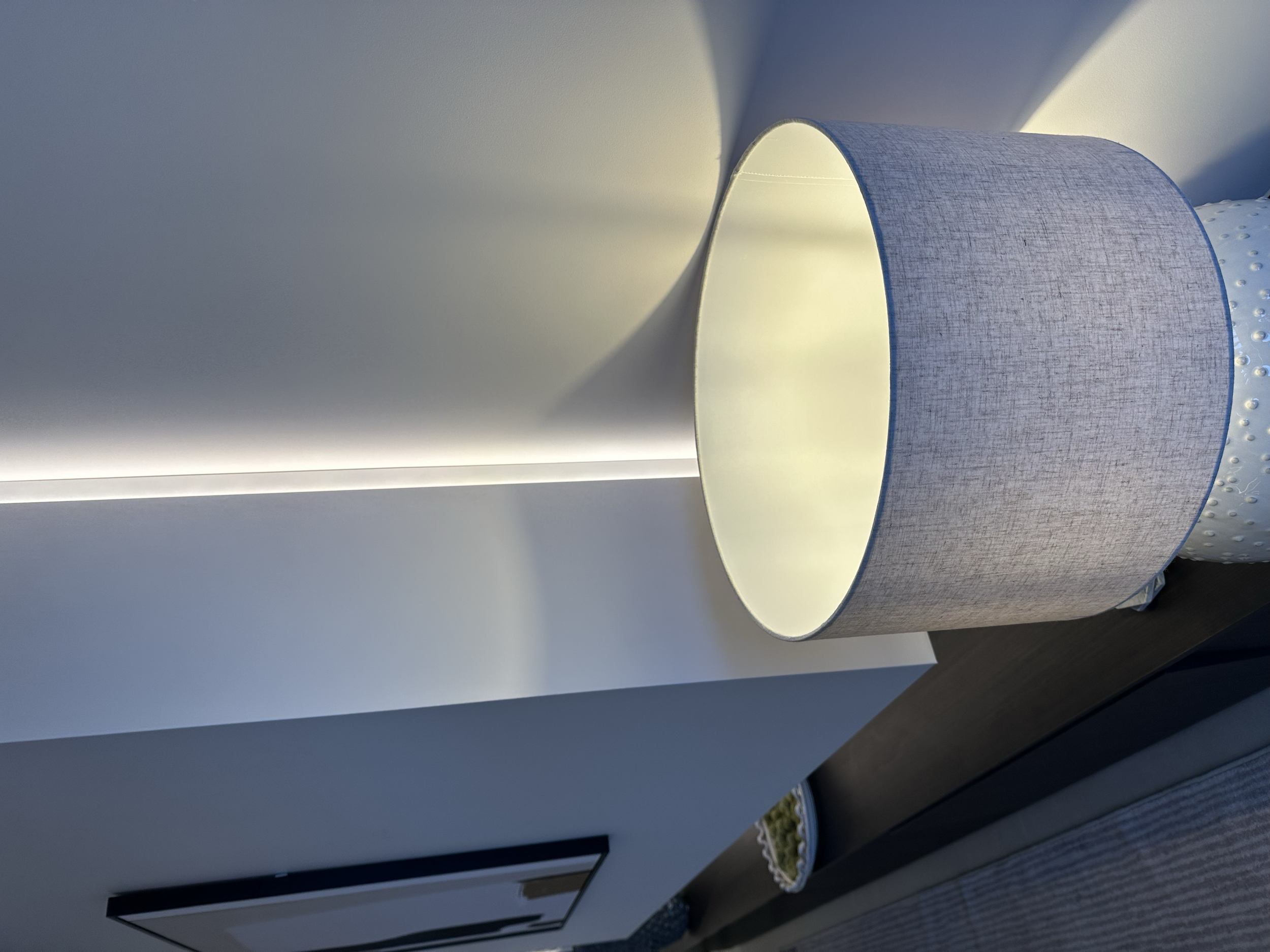 A visible gap between a floating media wall and the wall behind, in the foreground is a modern table lamp. The lamp is illuminated, casting light and shadows on the wall behind it.