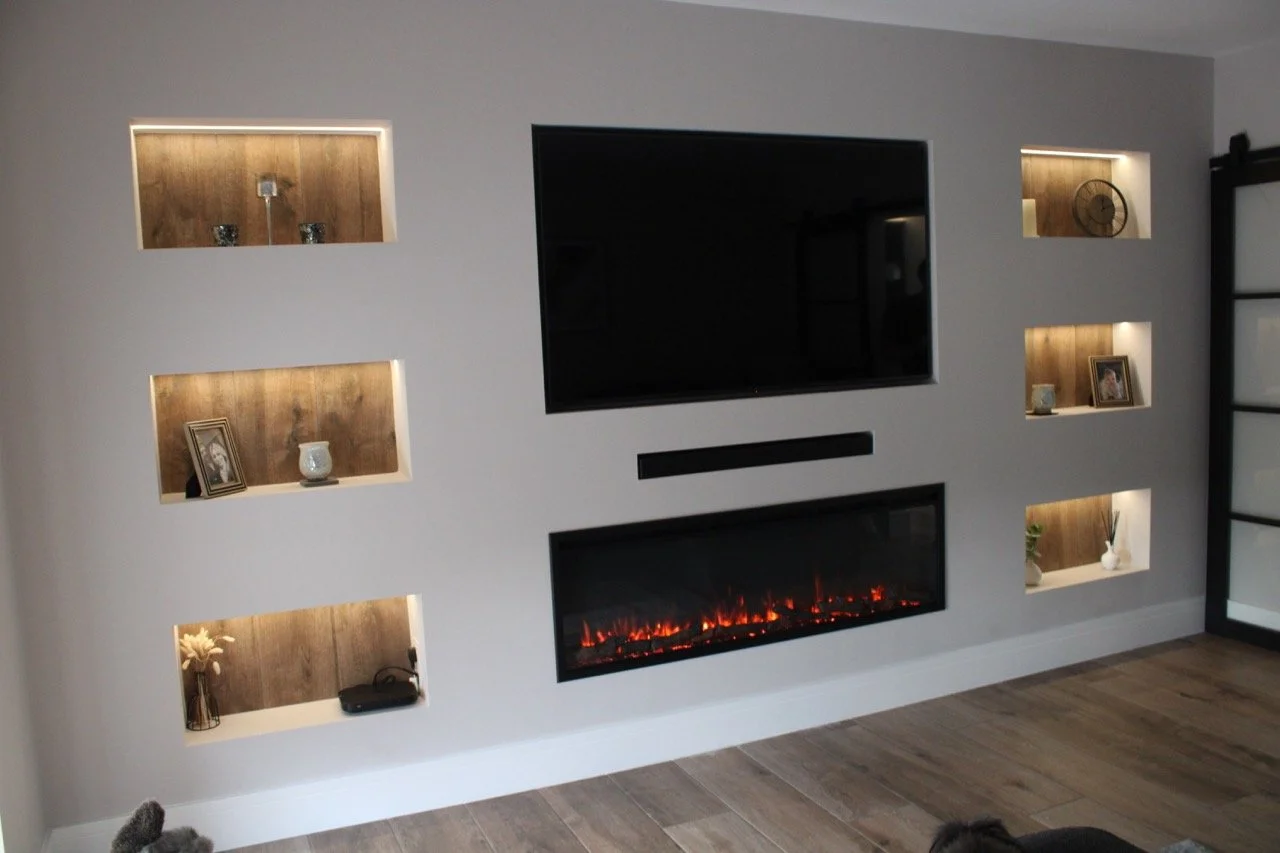 Modern living room wall with built-in shelves illuminated by lighting, a television above an integrated electric fireplace, and decorative items such as photo frames, vases, and a clock in the illuminated alcove units.