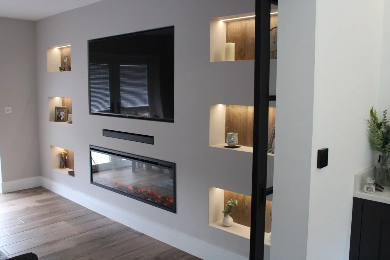 Living room with a modern media wall featuring a mounted flat-screen TV and a recessed electric fireplace, with illuminated built-in shelves displaying decorative items. Concealed cable management. 
