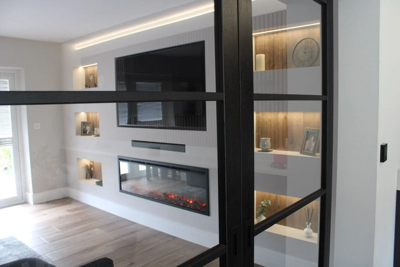 Modern living room with a built-in electric fireplace, wall-mounted TV, and illuminated wall alcoves with decorative items. Glass sliding door on the left, light wood flooring, and a black metal and glass partition.