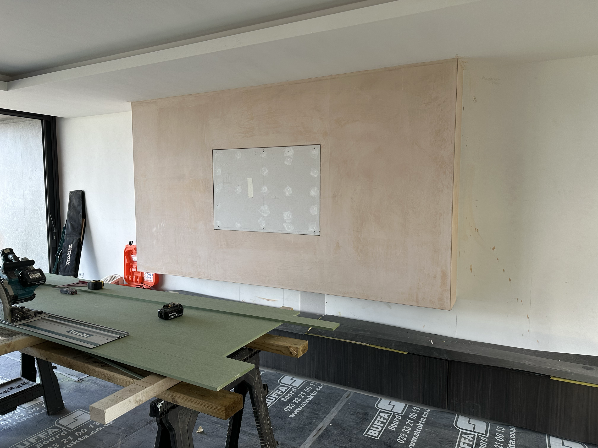 Interior of a media wall under construction with a floating TV wall, a window on the left, and work tools on a table in the foreground.