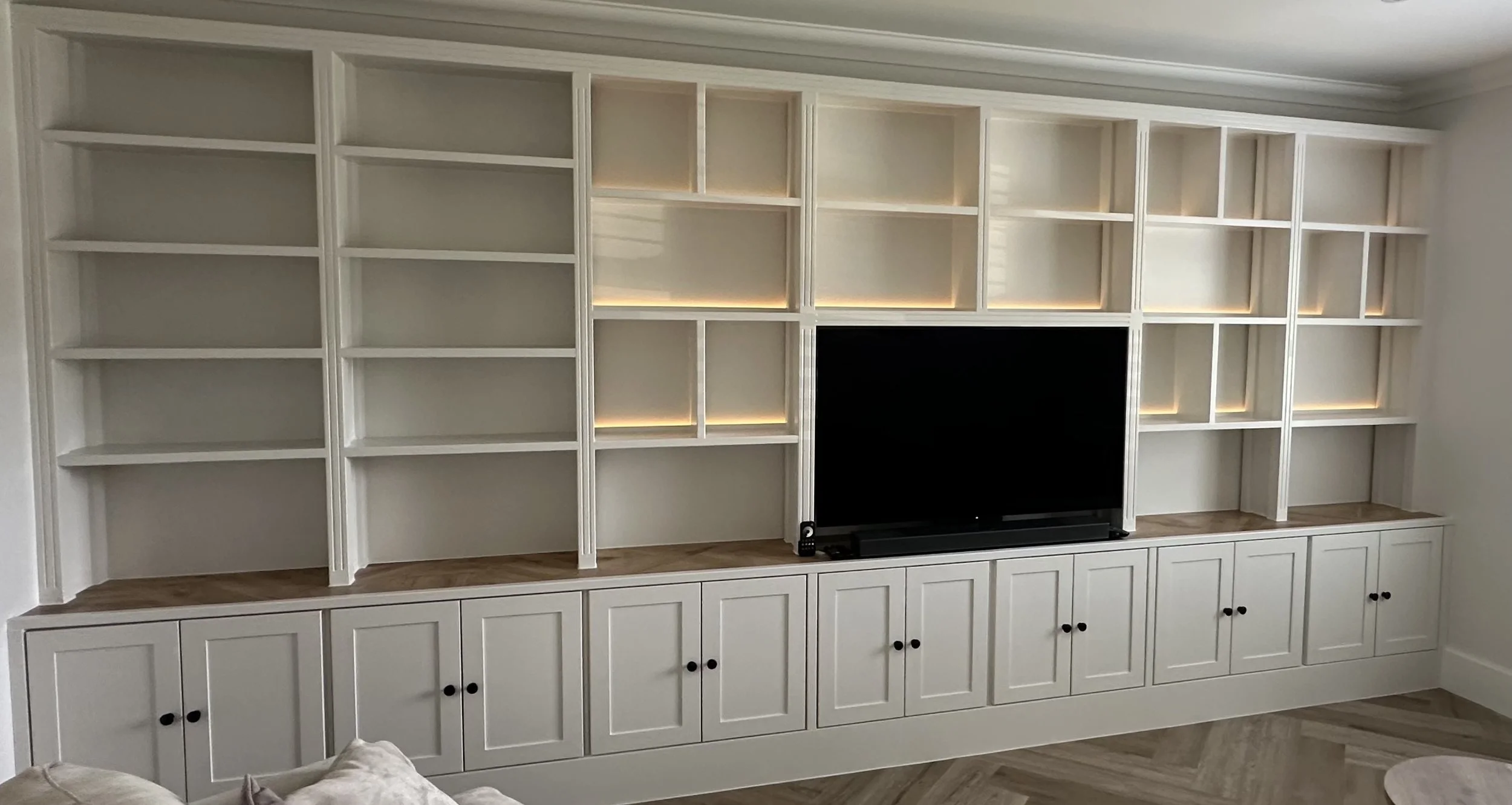 White built-in balanced shelving unit with multiple compartments for storage, some with LED lighting, and a central space for a TV, with cabinets at the bottom.