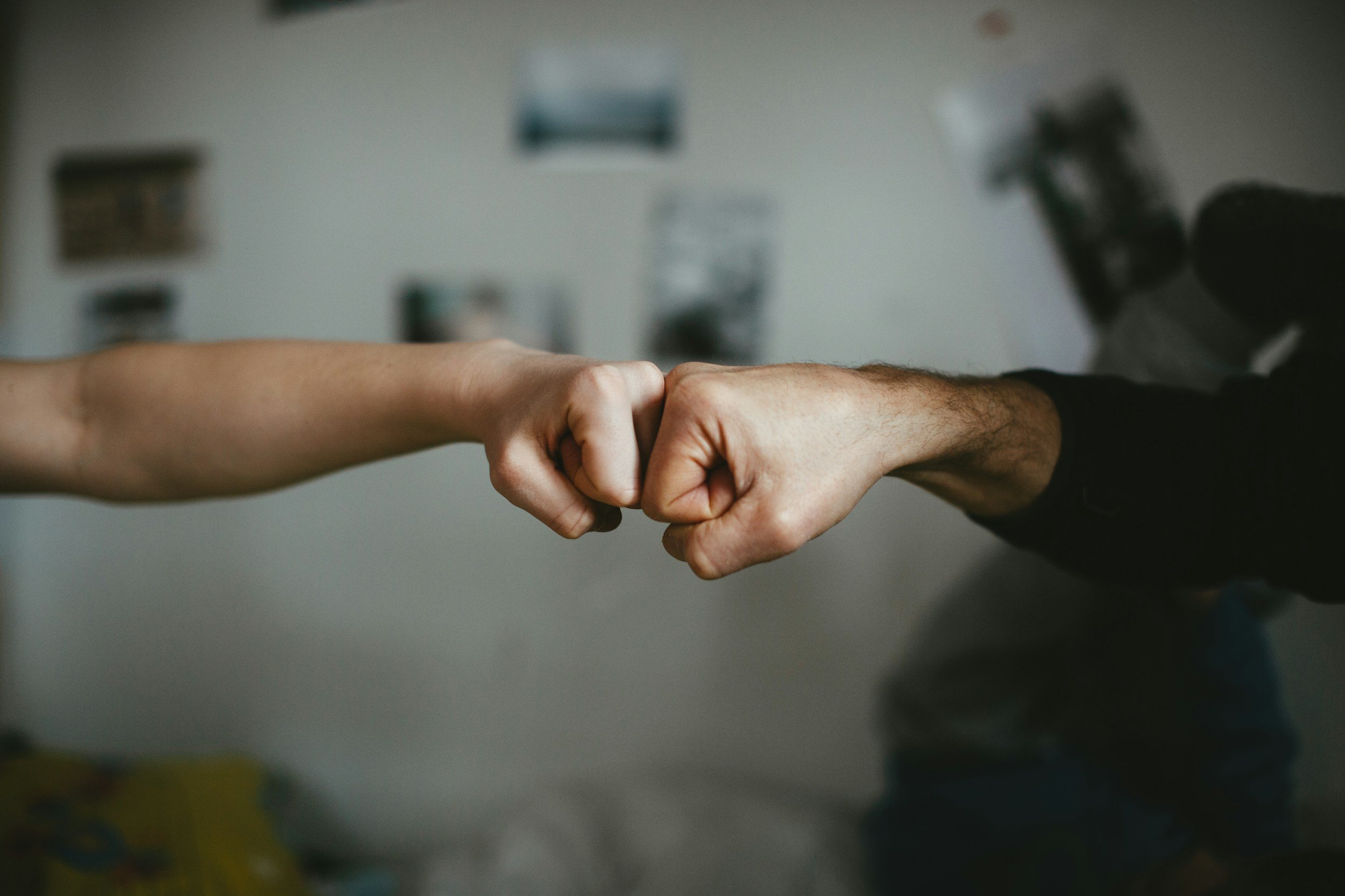 Close-up of two people fist bumping, one with a bare arm and the other with a black sleeve, in a room with blurred background.