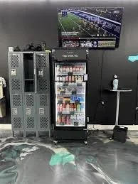 A vending machine in a room with lockers and a television mounted on the wall.