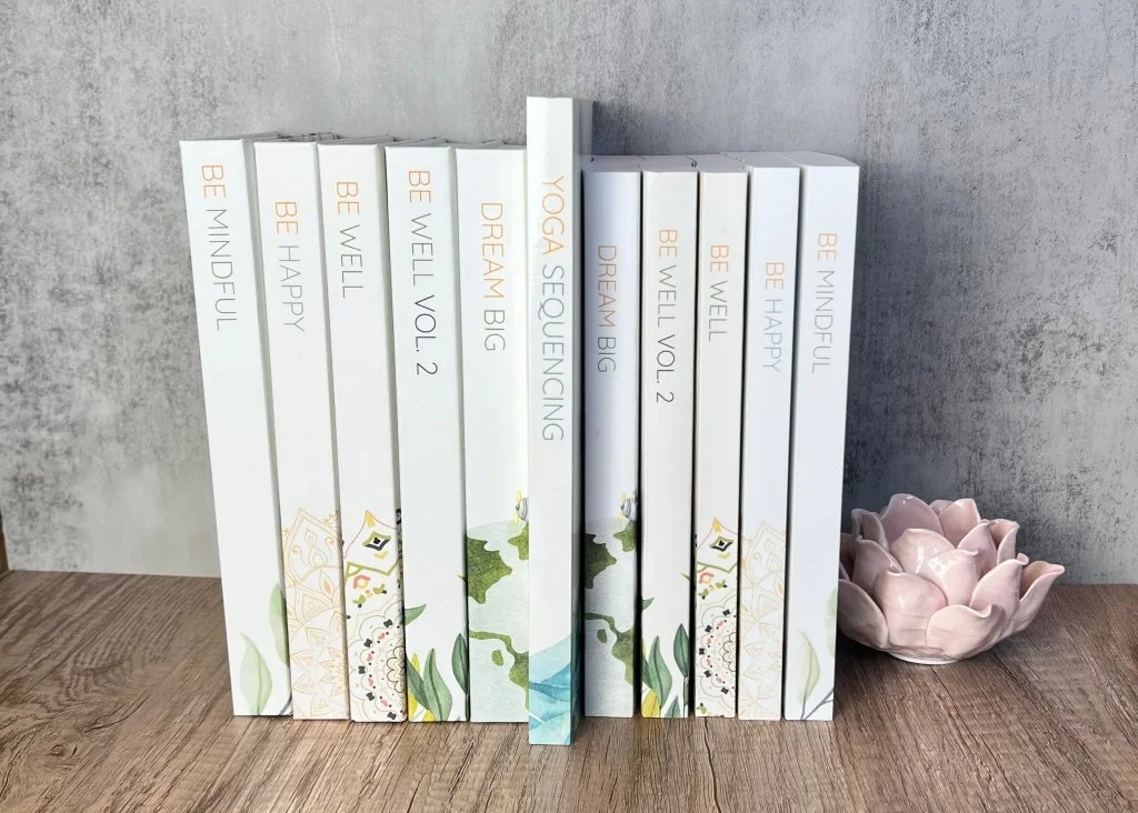 A collection of white books with colorful titles and designs, arranged on a wooden surface with a gray textured background, with a pink ceramic lotus flower bowl to the right.