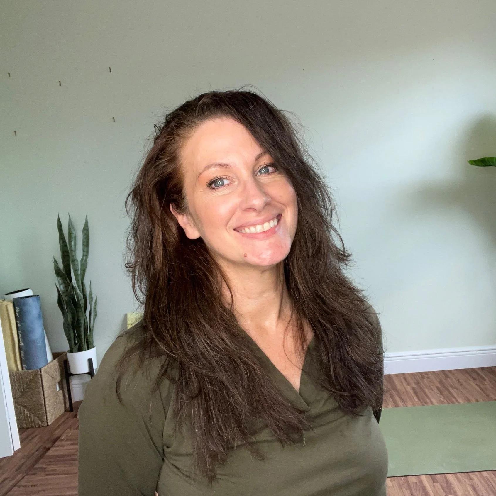 A woman with long, wavy brown hair smiling in a room with green walls, a potted plant, and books in the background.