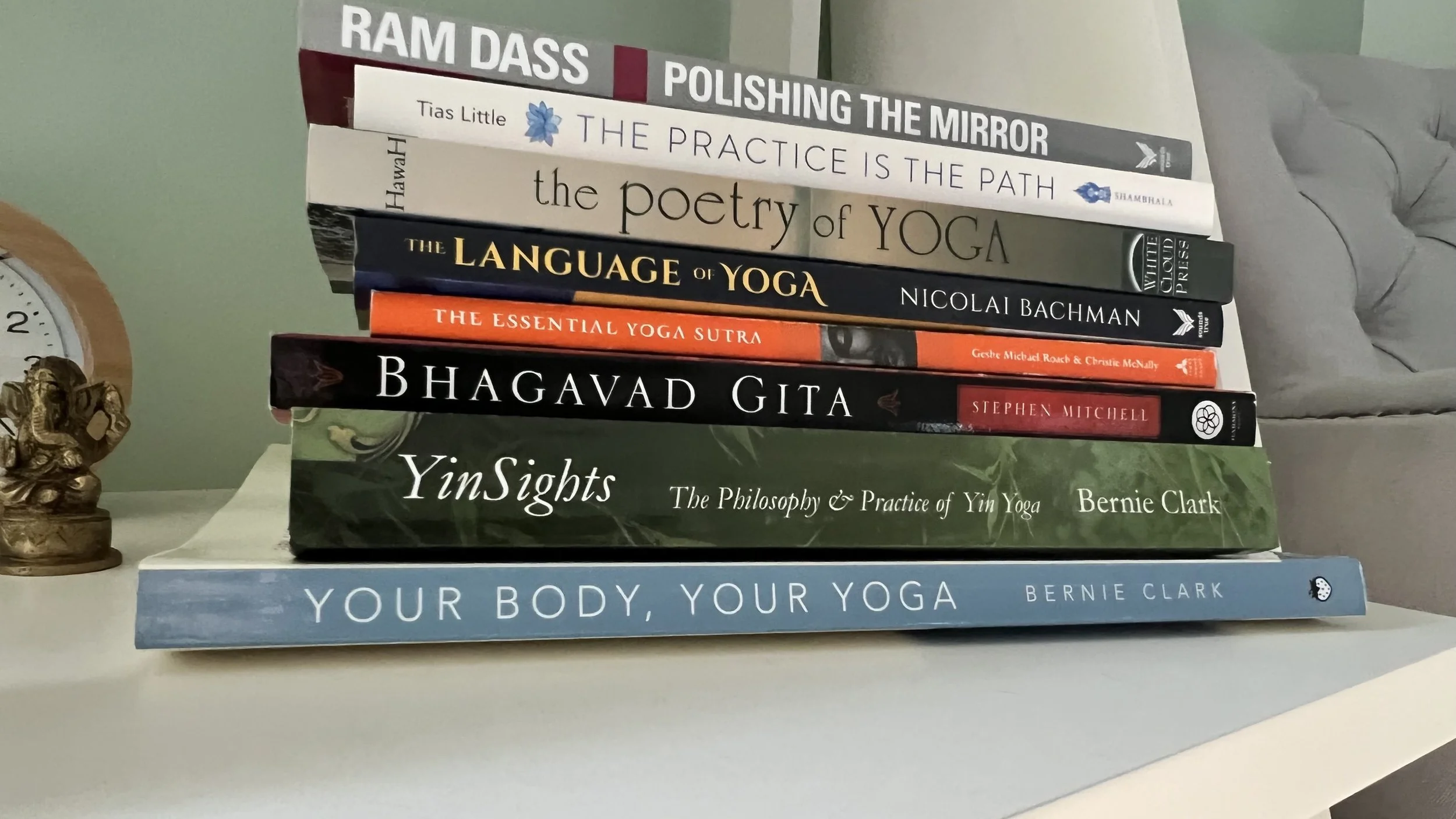Stack of eight books on a white surface, with titles related to yoga and philosophy, including 'Ram Dass Polishing the Mirror', 'The Practice is the Path', 'The Poetry of Yoga', 'The Language of Yoga', 'The Essential Yoga Sutra', 'Bhagavad Gita', 'Yin Sights', and 'Your Body, Your Yoga'. A small statue of Ganesha and a clock are to the left, and a gray cushion is in the background.