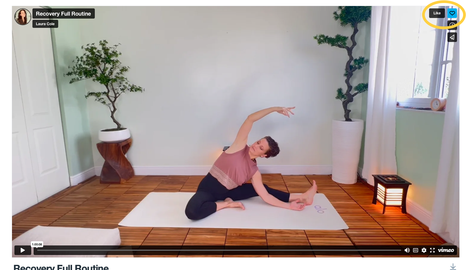 A woman practicing yoga on a white mat in a room with wooden flooring and light green walls, with tall potted plants and a window with white curtains. She is wearing a pink top and black pants, seated in a side split pose with one arm extended over her head and the other reaching toward her foot.
