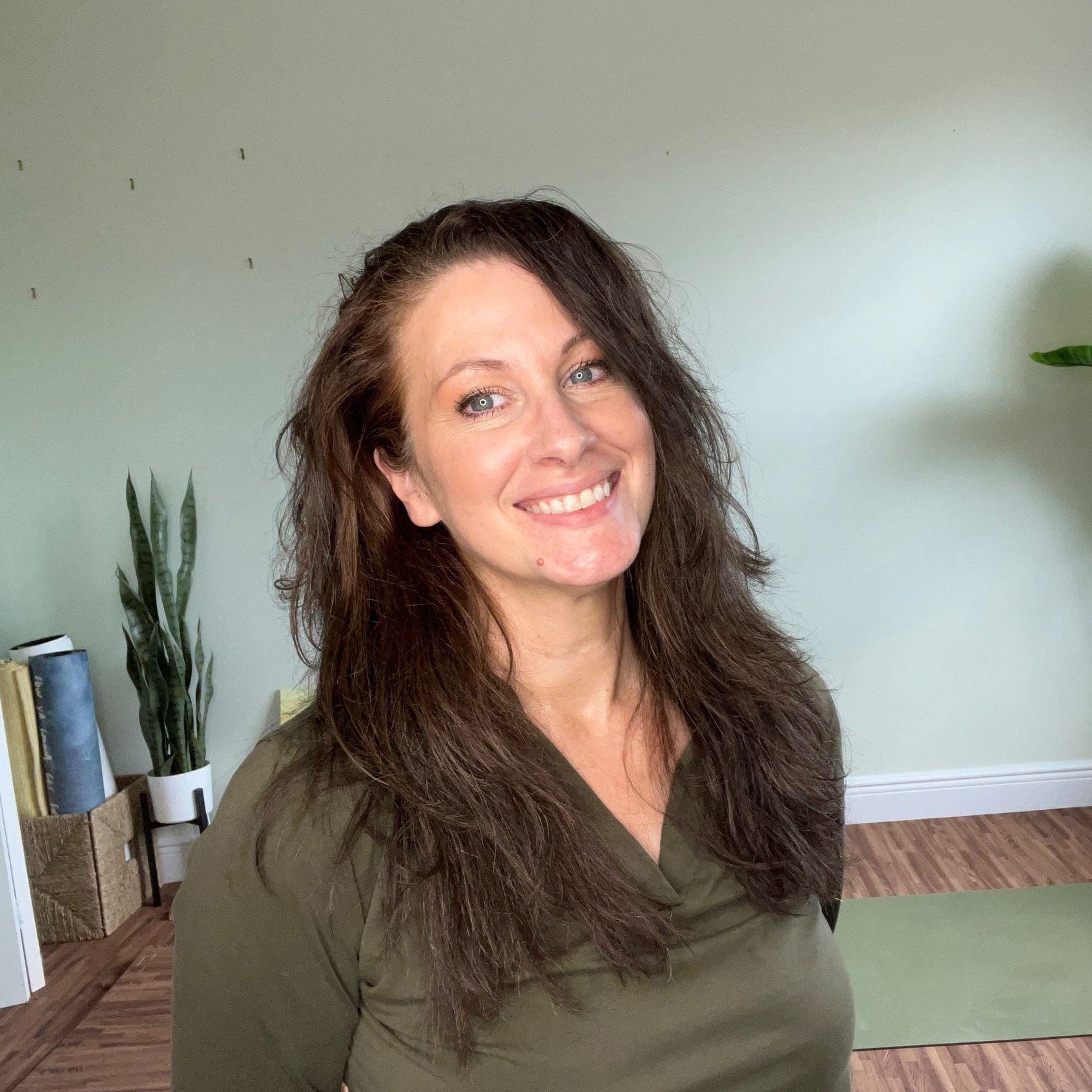 A woman with long, wavy brown hair smiling indoors near a light green wall with a potted plant and books in the background.