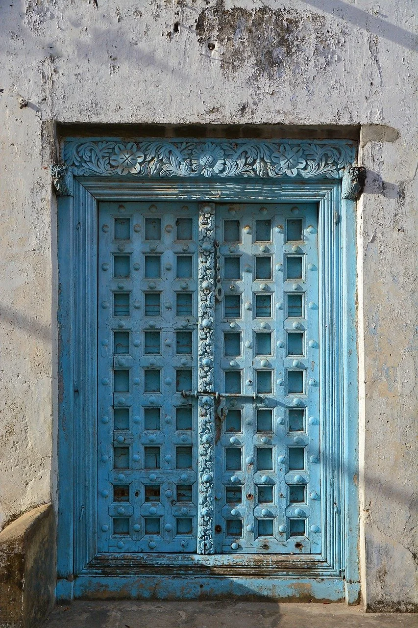 Blue wooden door with intricate carvings and square patterns, set in a weathered white wall evocative of locations found on the Swahili Coast such as Zanzibar and Lamu Island.