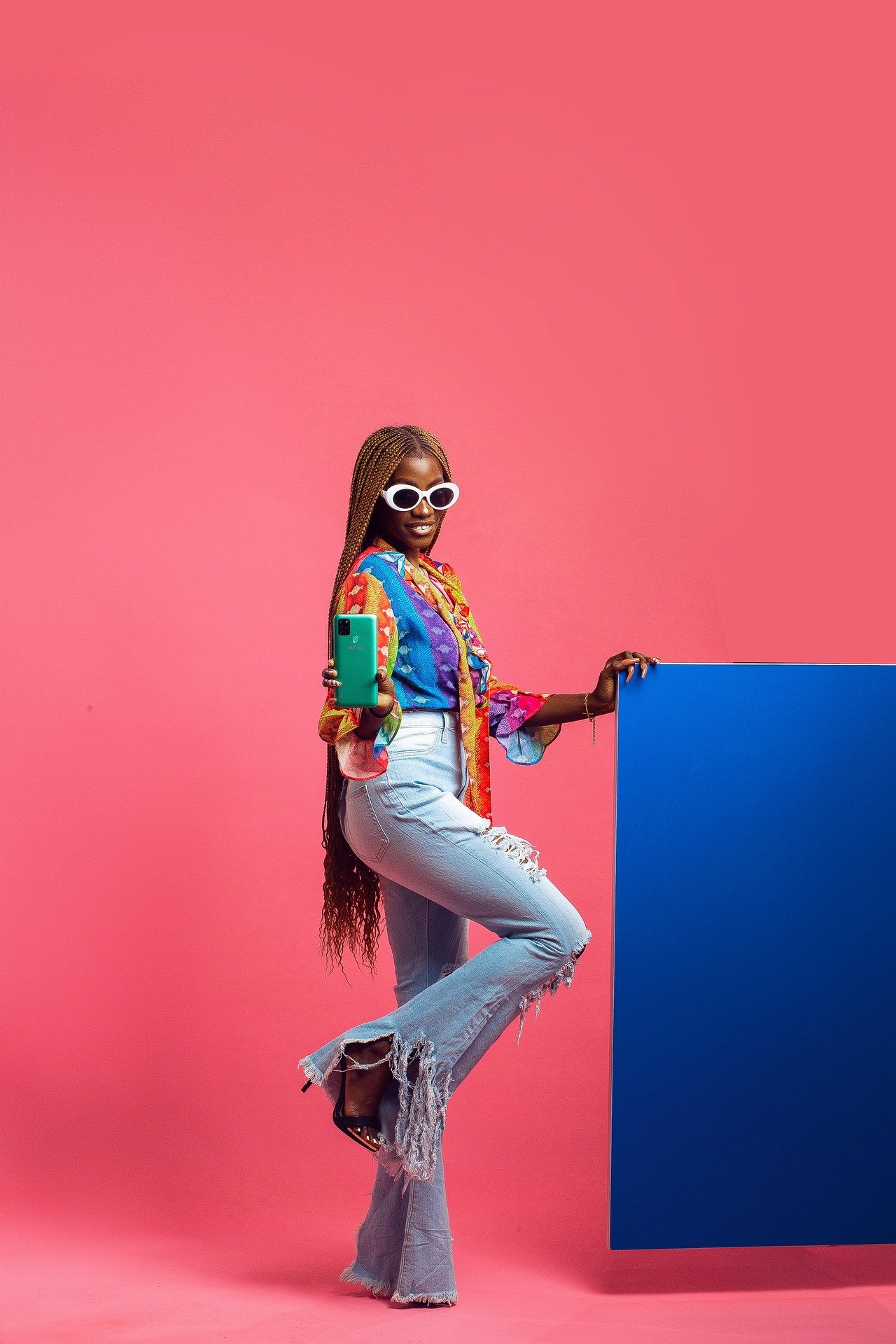 A woman with long braided hair wearing large white sunglasses, a colorful patterned shirt, and distressed jeans, taking a selfie with a green smartphone against a pink background with a blue panel.