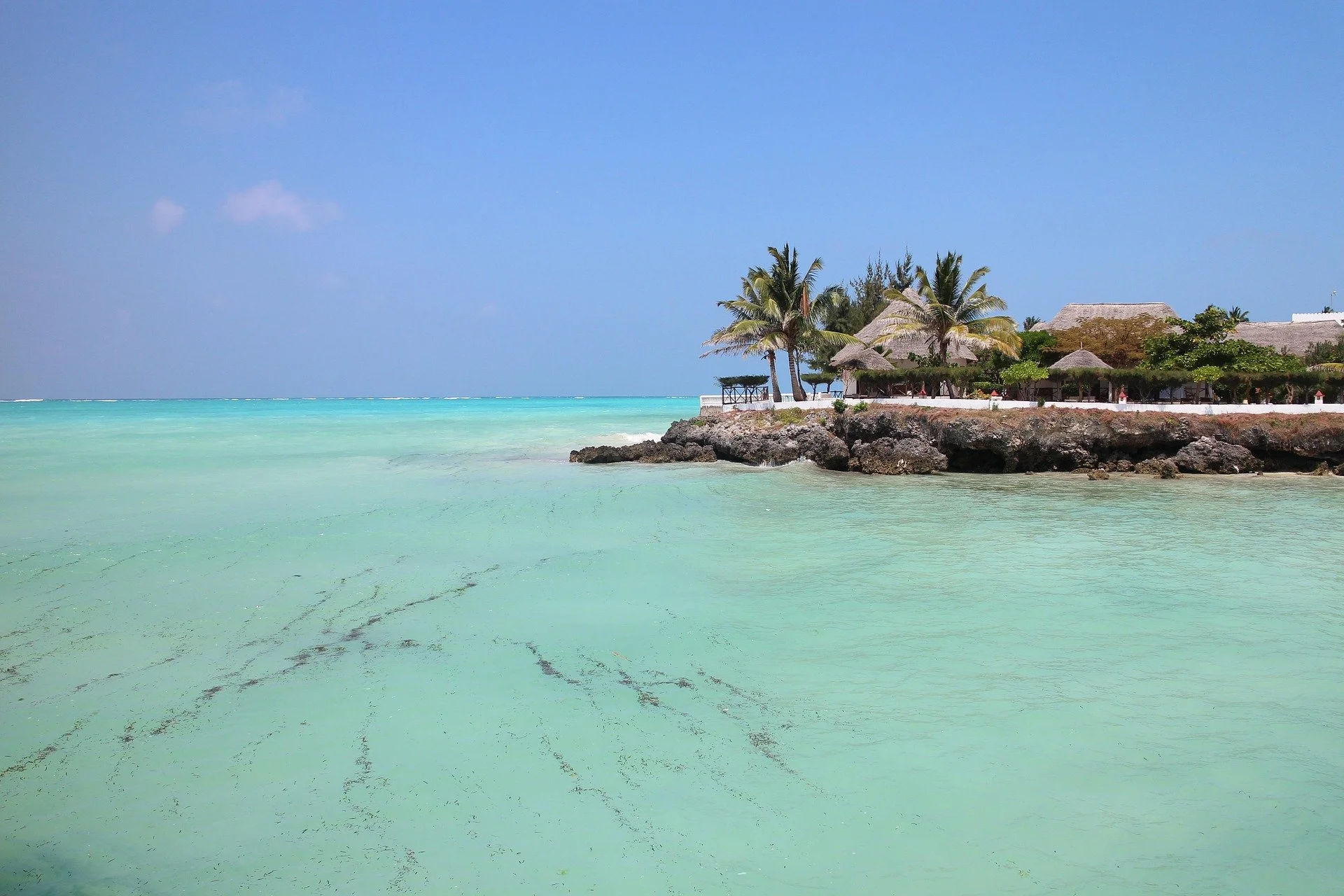 Tropical beach with turquoise water, rocky shoreline, palm trees, and thatched-roof structures under a clear blue sky.