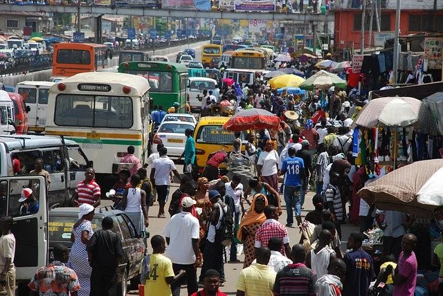 A busy street market in Ghana with many pedestrians, colorful umbrellas, and various vehicles including buses and taxis.
