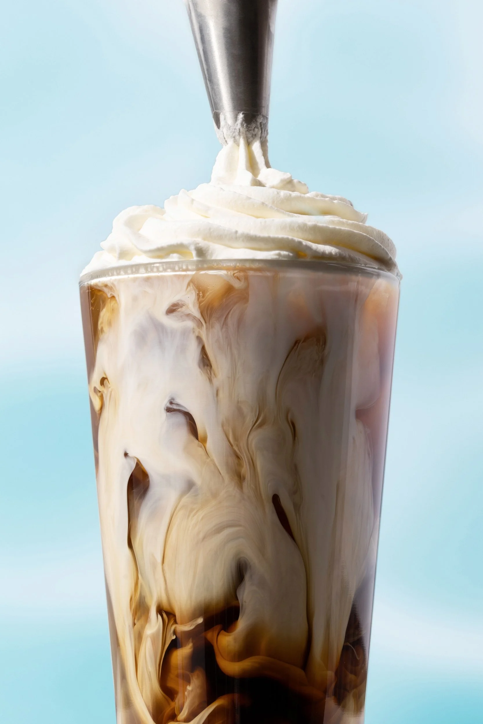 Close-up of a tall glass of iced coffee with swirling cream, topped with whipped cream, and a straw.
