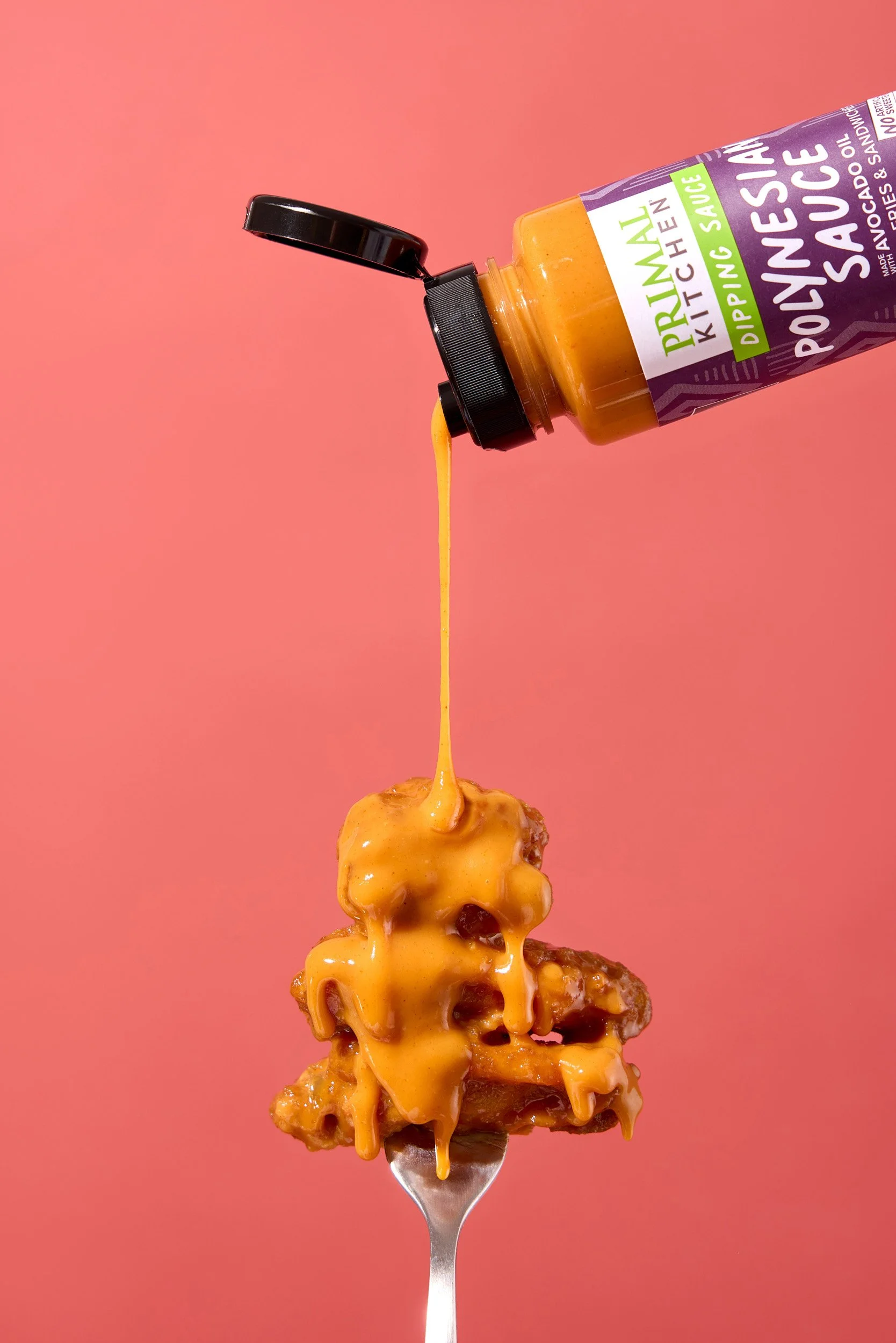 A fork with fried chicken covered in spicy mayonnaise is being drizzled with yellow mustard from a bottle labeled 'Primal Kitchen Sriracha.'