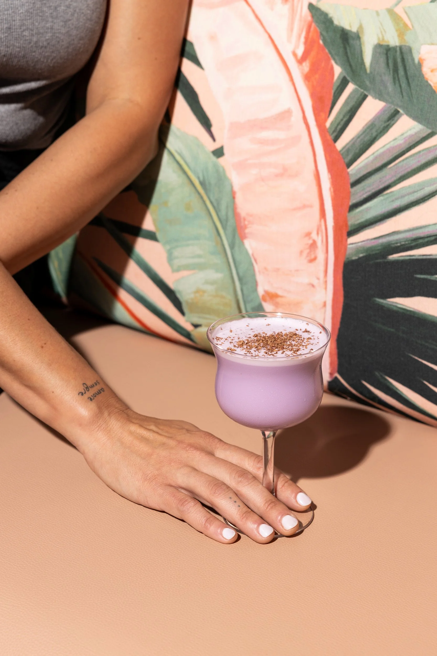 A woman with a tattoo on her wrist resting her hand on a beige surface, holding a purple cocktail glass with a frothy pink drink sprinkled with herbs, in front of a tropical leaf-patterned background.