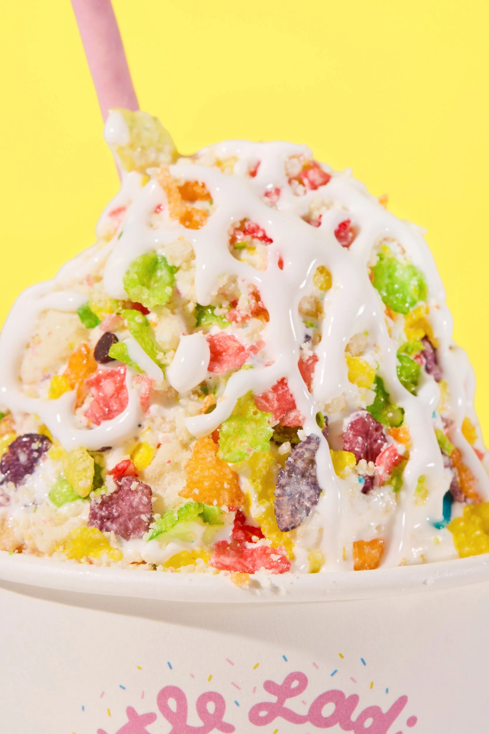 Close-up of colorful ice cream with sprinkles and white chocolate drizzle in a cup, yellow background.