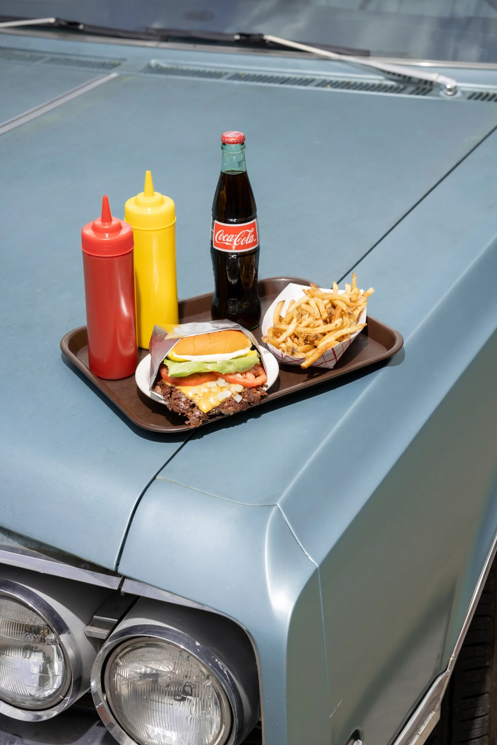 Tray with a hamburger, fries, a bottle of Coca-Cola, and condiments on the hood of a vintage blue car.