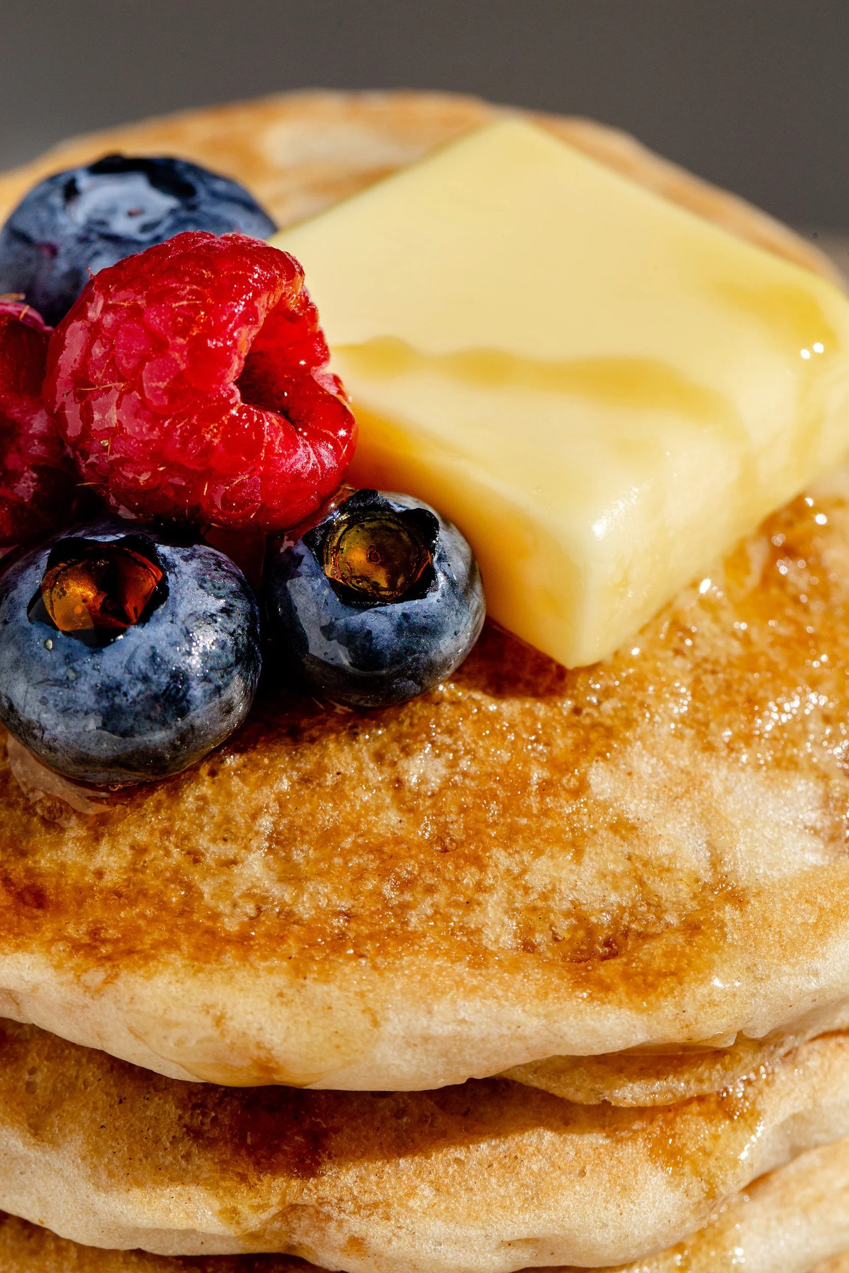Close-up of a stack of pancakes topped with a pat of butter, fresh blueberries, raspberries, and syrup.