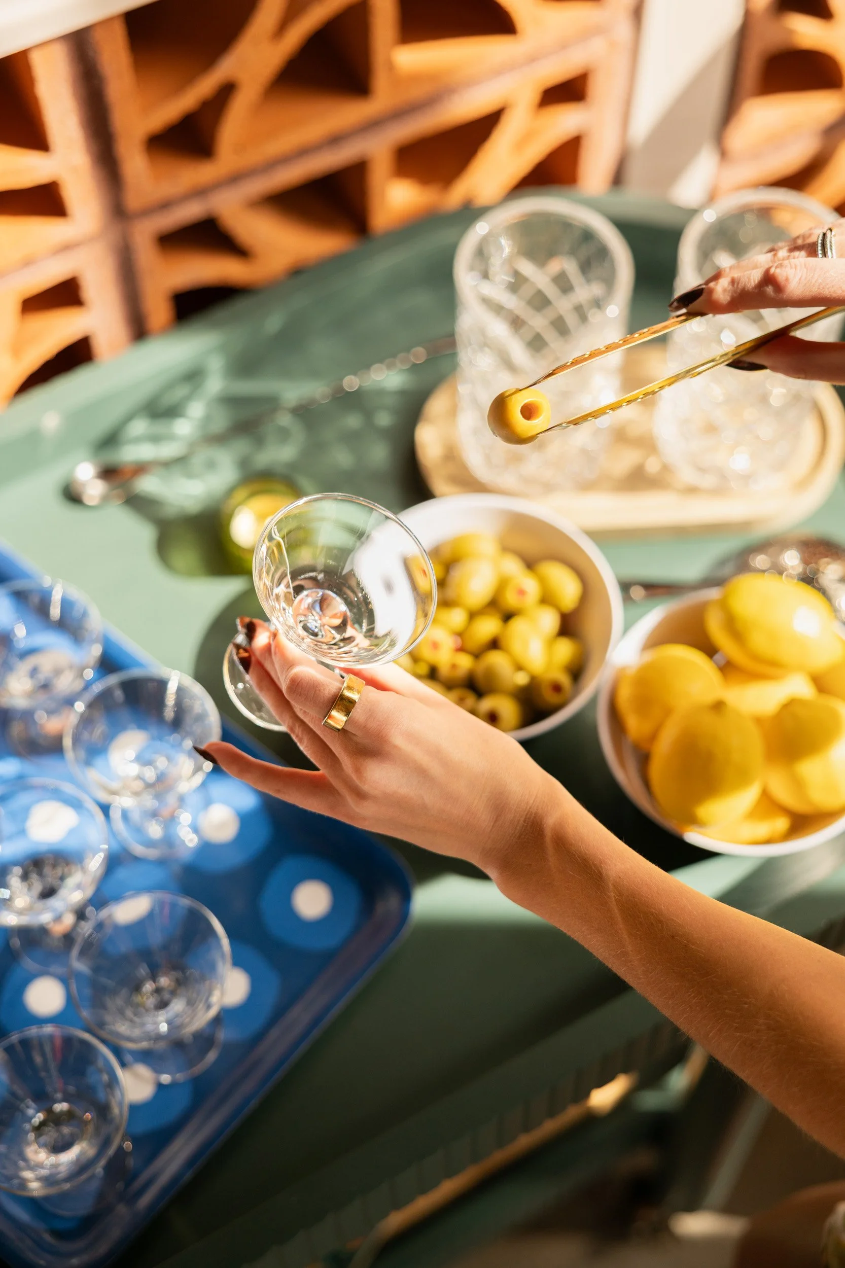 A person is holding a clear glass in one hand and tongs with a green olive in the other, preparing to add it to the glass. The table features bowls of green olives and yellow lemons, a blue tray with empty glasses, and a decorative wooden backdrop.