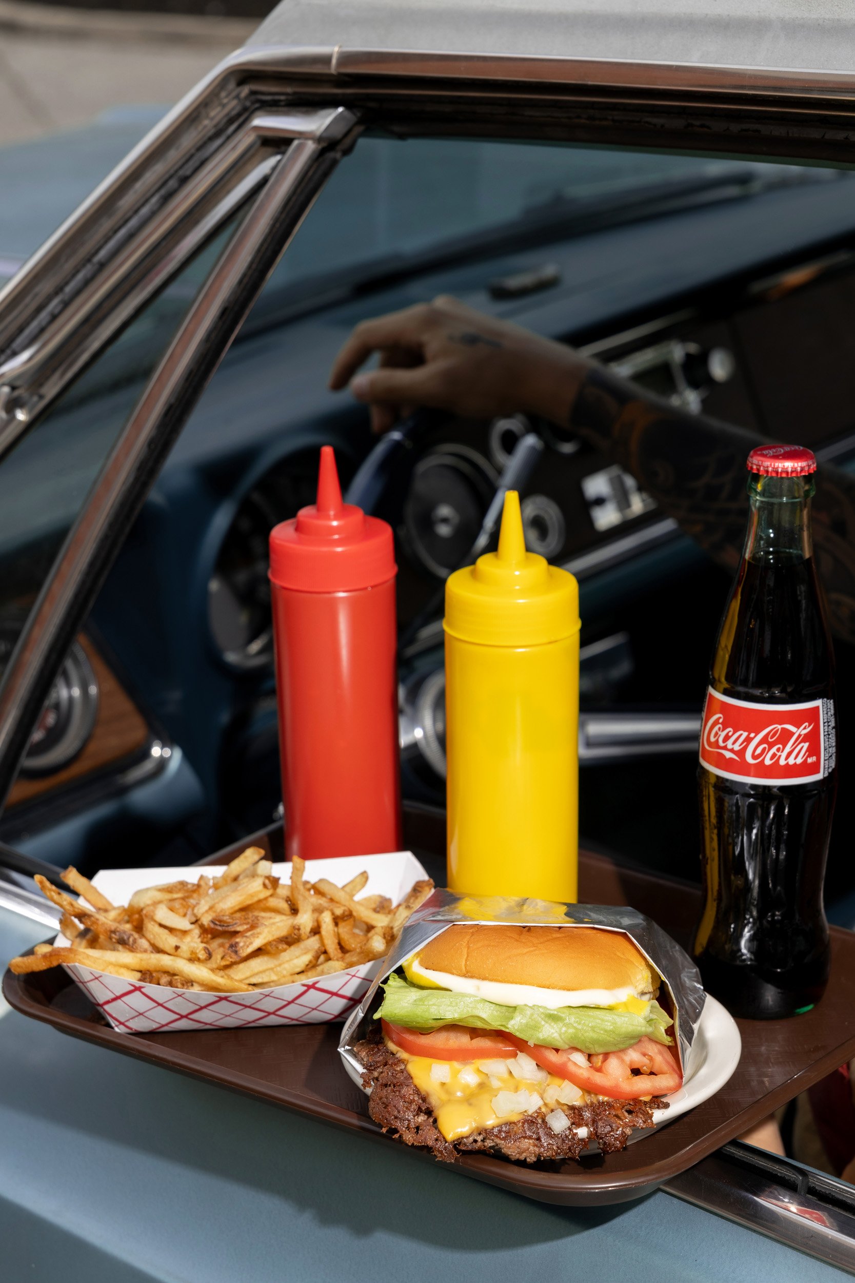 A tray of food in a vintage car shows a burger with lettuce, tomato, cheese, onions, and a beef patty, a serving of French fries, ketchup and mustard bottles, and a bottle of Coca-Cola.