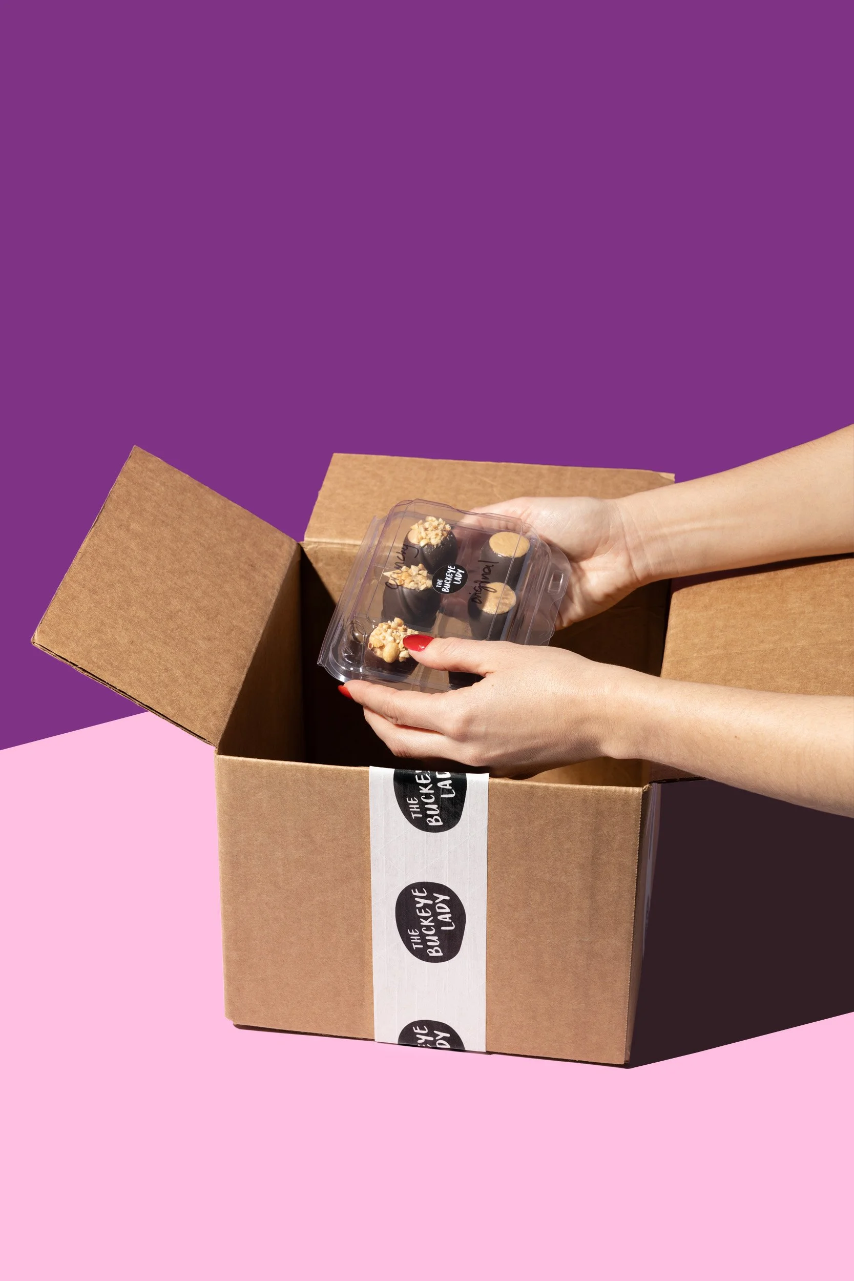 A person with red painted nails holding a container of chocolate desserts with toppings, about to be placed in a cardboard box with a black and white 'The Buckeye Lady' logo, against a purple and pink background.