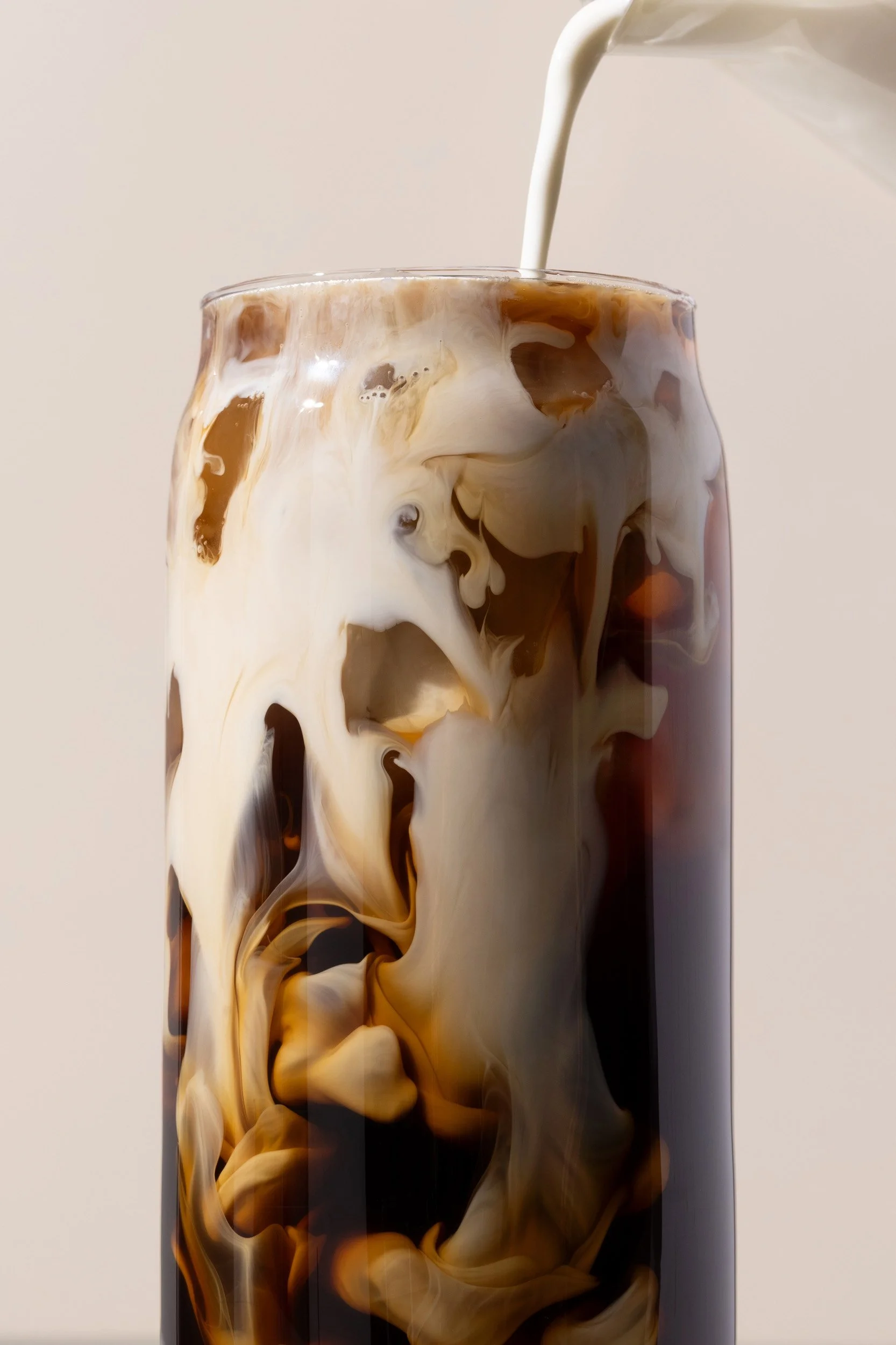A tall glass of iced coffee with milk being poured in, creating swirling patterns.