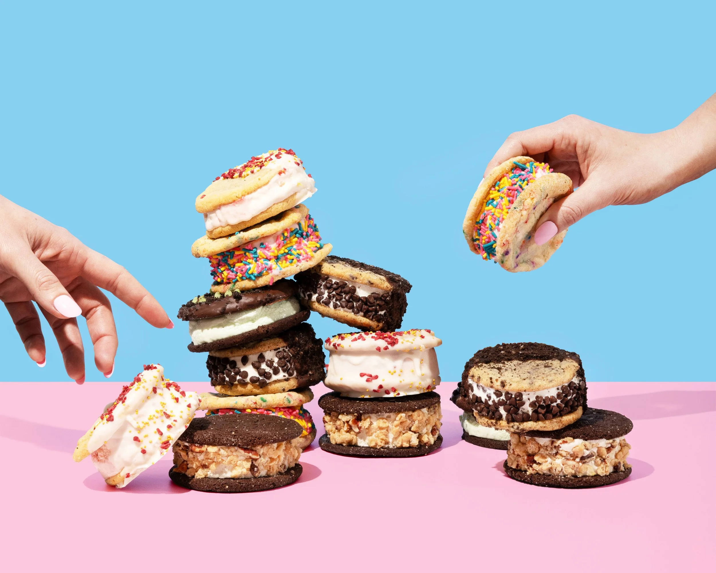 Hands placing and reaching for a variety of colorful cookies and whoopie pies on a pink and blue background.