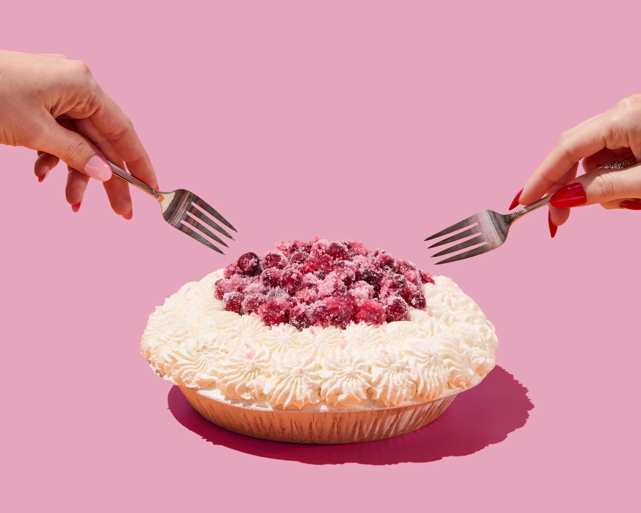 A dessert topped with whipped cream and berries, with two hands holding forks facing each other against a pink background.