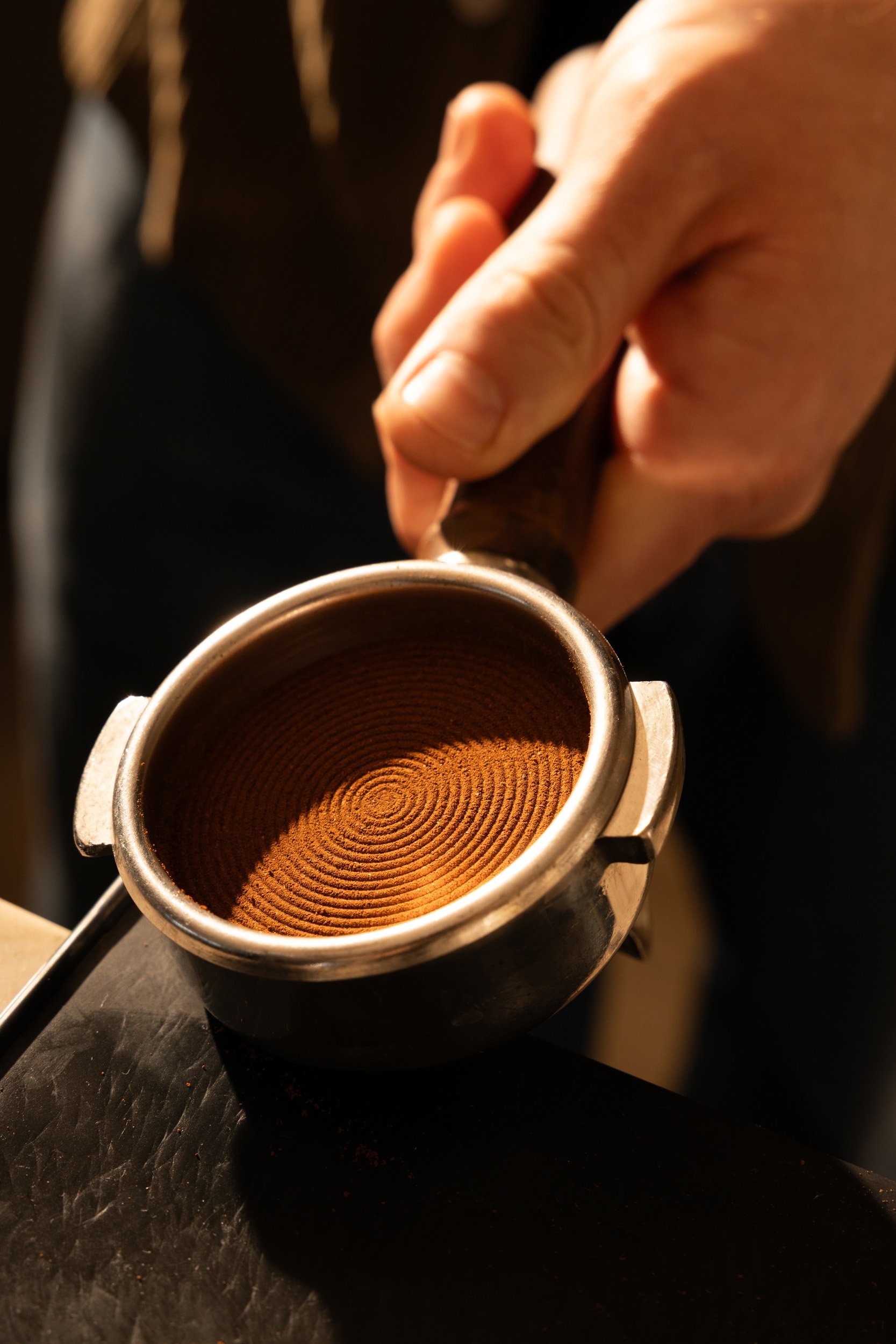 A hand holds a coffee tamper over a portafilter with freshly tamped espresso grounds.