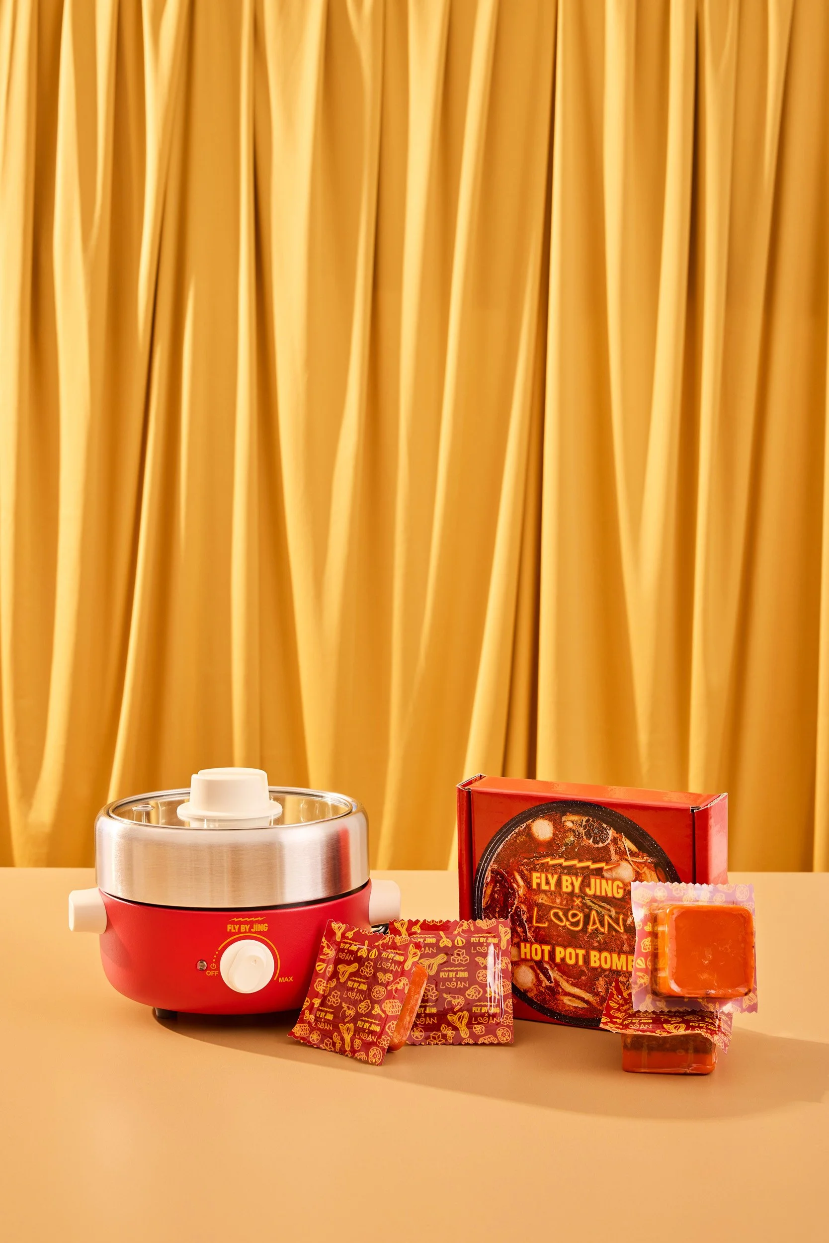 A hot pot stove with two small packets, a box of hot pot bombs, and a small bar of soap or caramel, set against a yellow curtain background.