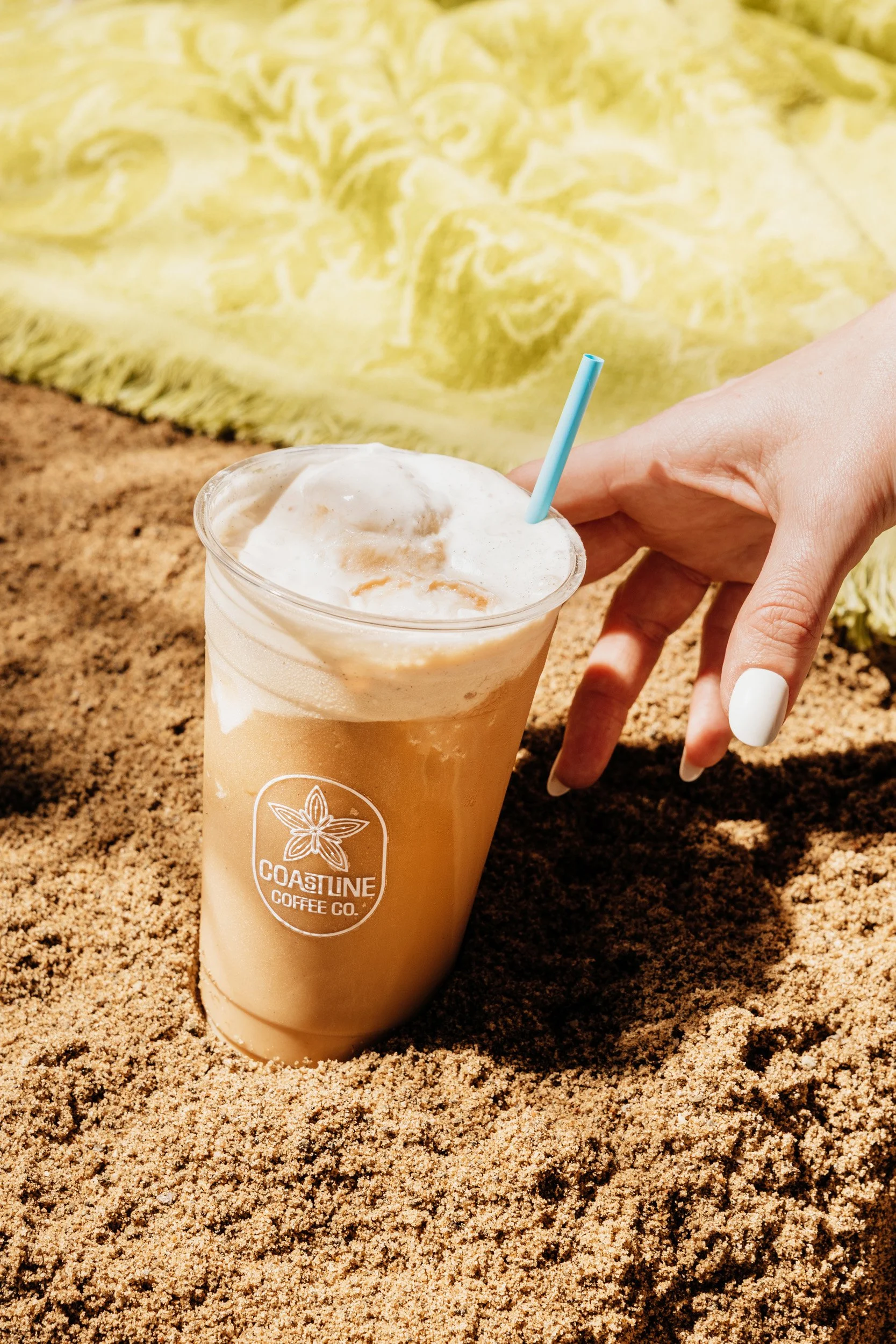 A hand with white painted nails reaching for a blended coffee drink in a clear plastic cup with a straw, placed on sandy ground with a yellow towel in the background. The cup has a logo for Coastline Coffee Co.