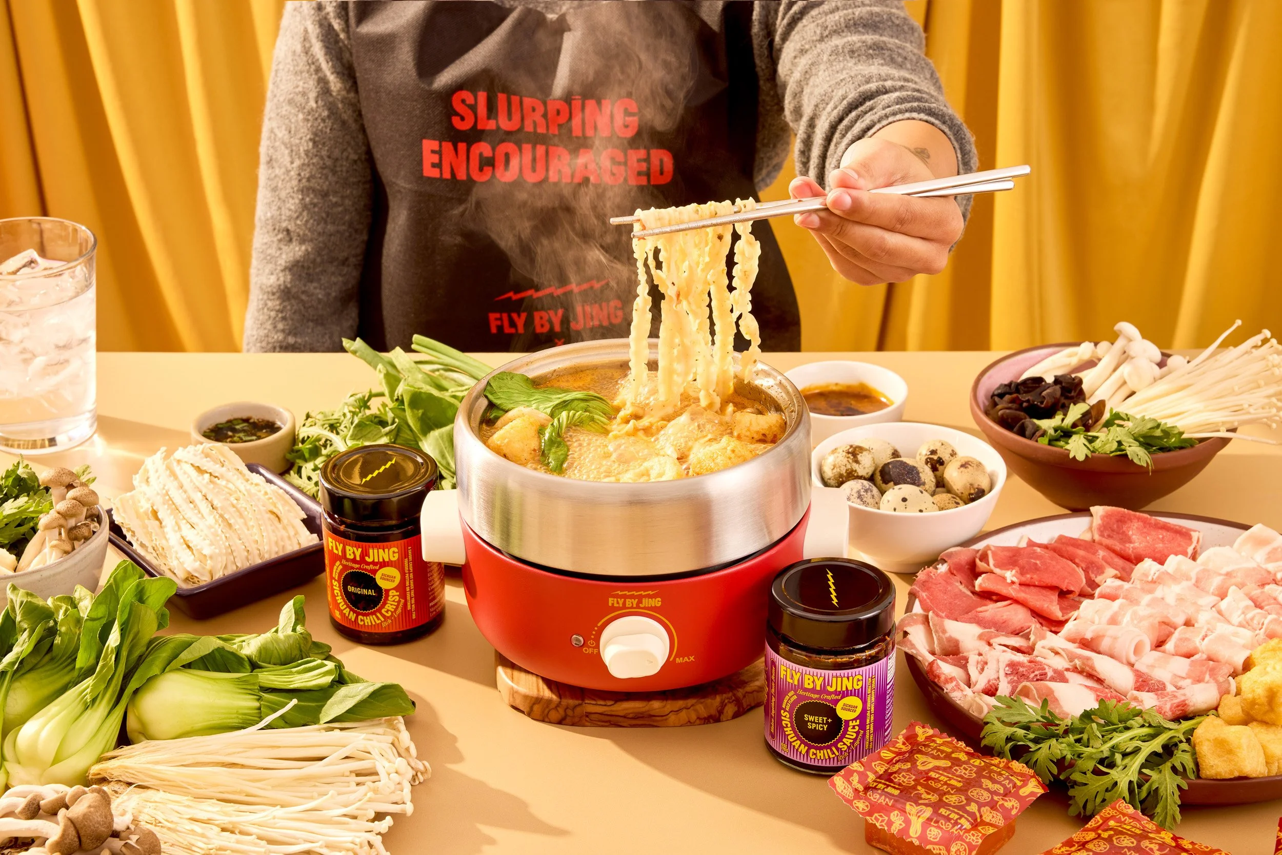 Person holding chopsticks lifting noodles from a hot pot surrounded by vegetables, meats, sauces, and condiments on a dining table.