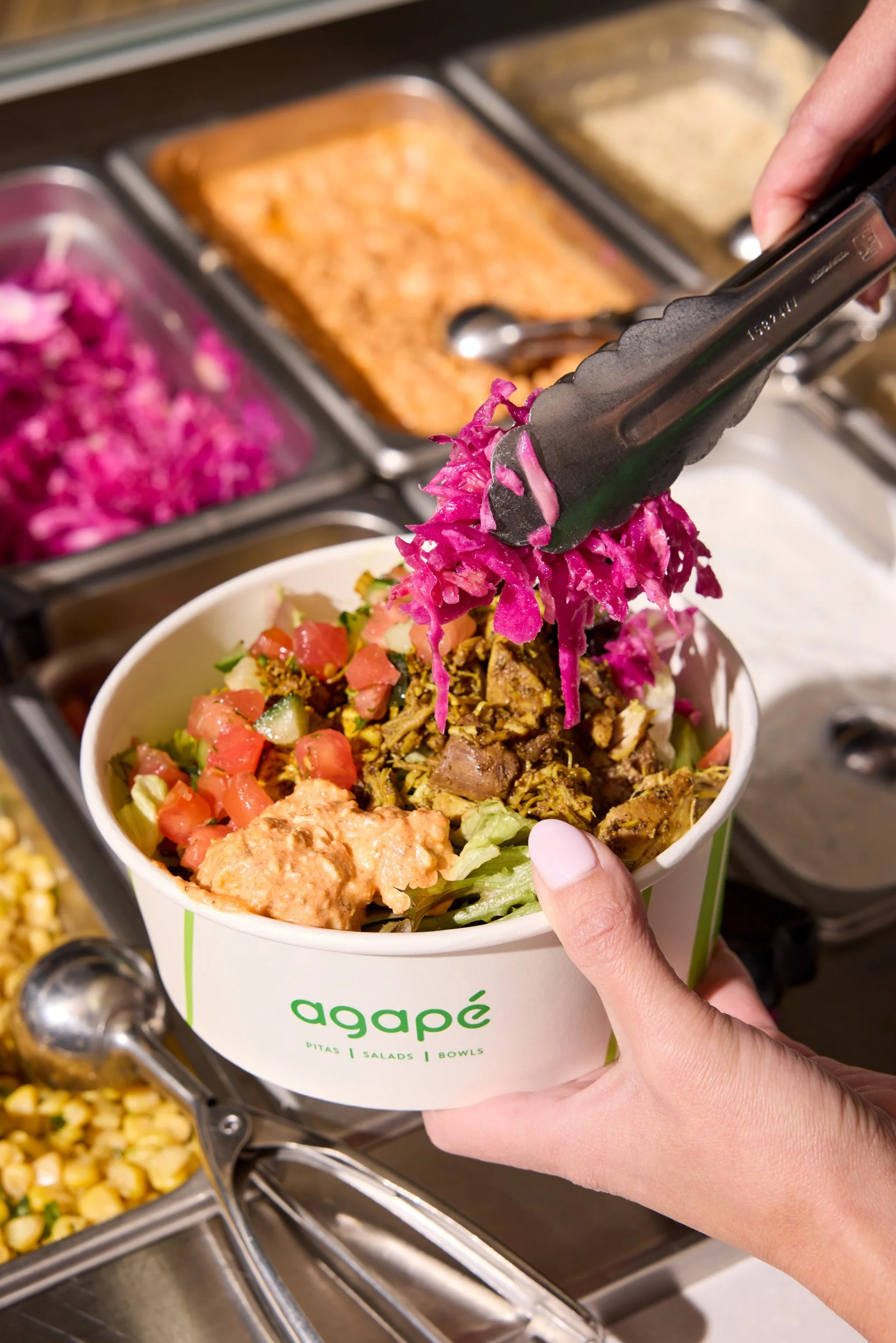 Person adding shredded pink cabbage to a salad in a white bowl labeled 'agapè' at a buffet line with various food trays in the background.