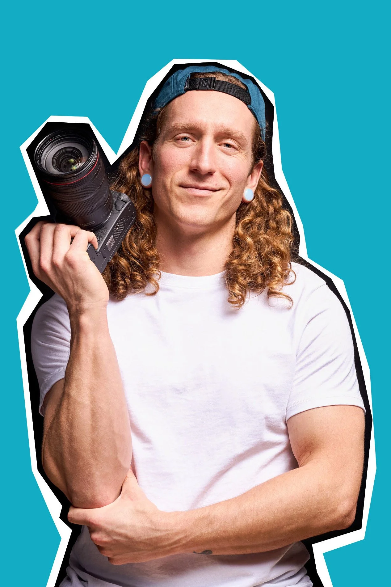 A young man with long curly hair, light skin, a backward cap, and earrings, holding a camera on his shoulder with a slight smile.