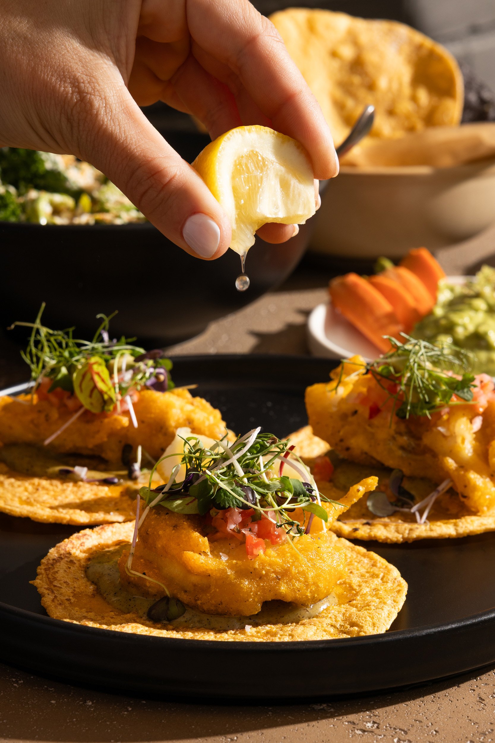 Person squeezing a lemon over fish tacos topped with microgreens, on a black plate.