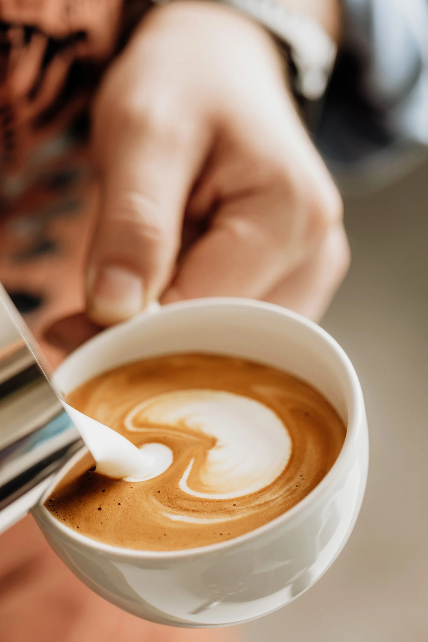 A person holding a white coffee cup with a latte art heart design on top, in a cozy setting.