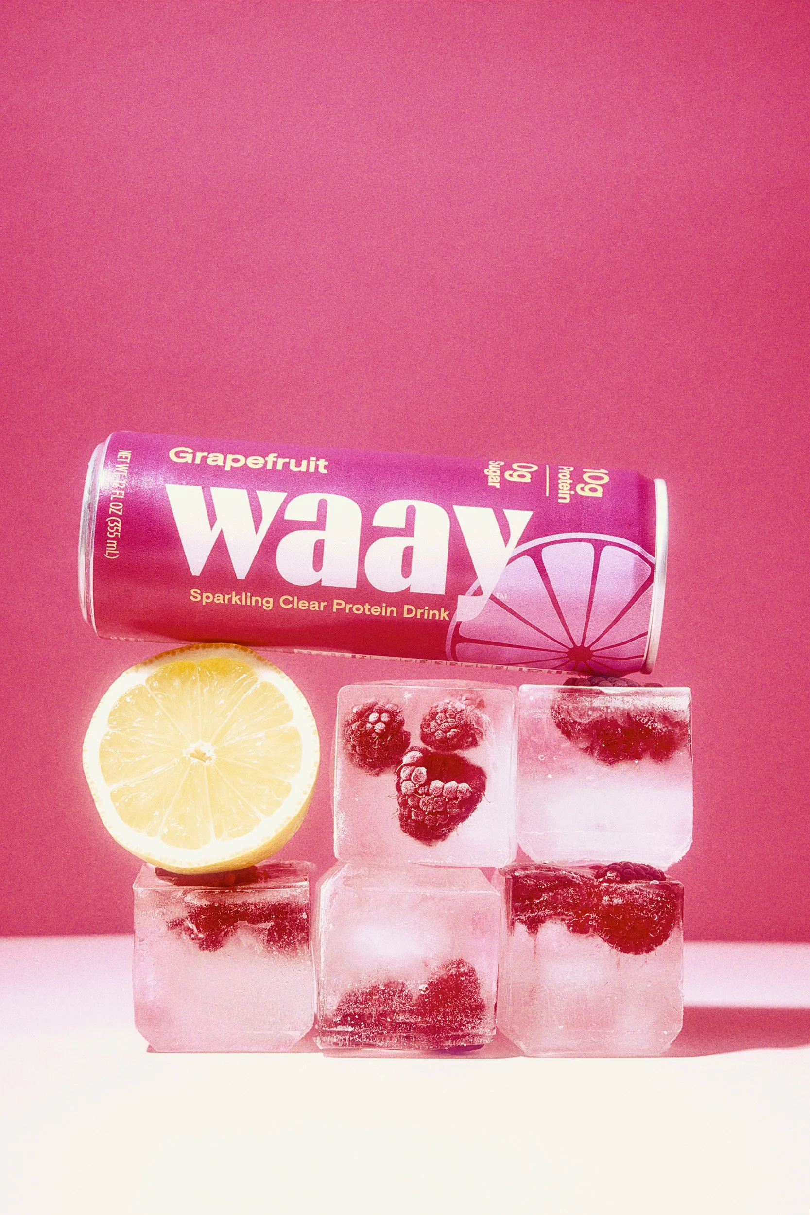 A pink can of Wawy sparkling clear protein drink with grapefruit flavor, a lemon half, and six ice cubes with berries inside, set against a pink background.