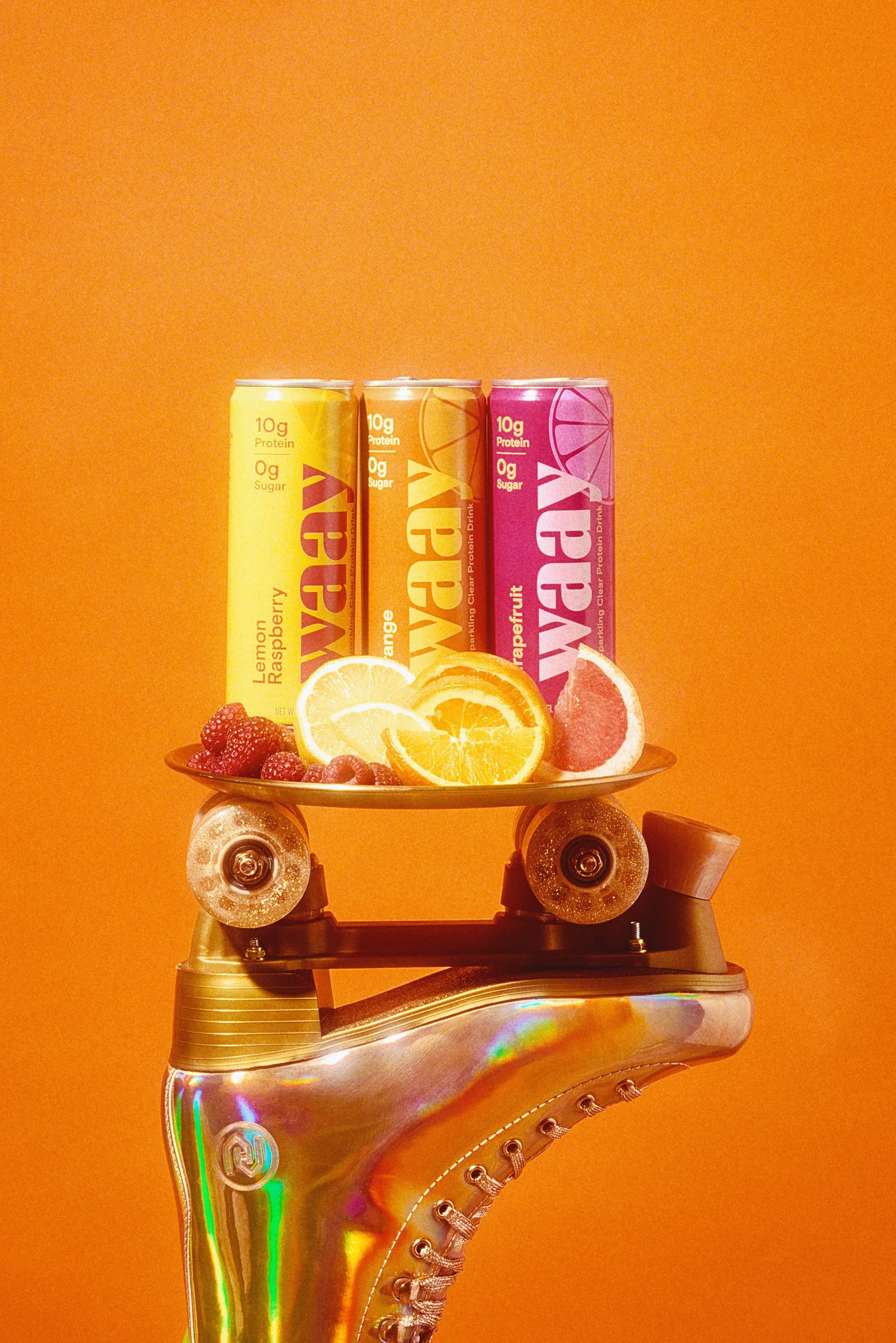 An ice skate with a gold finish features a skateboard deck with fruit and three colorful cans of flavored water on top of the skate. The fruits include lemon, orange, grapefruit, and raspberries, set against a bright orange background.