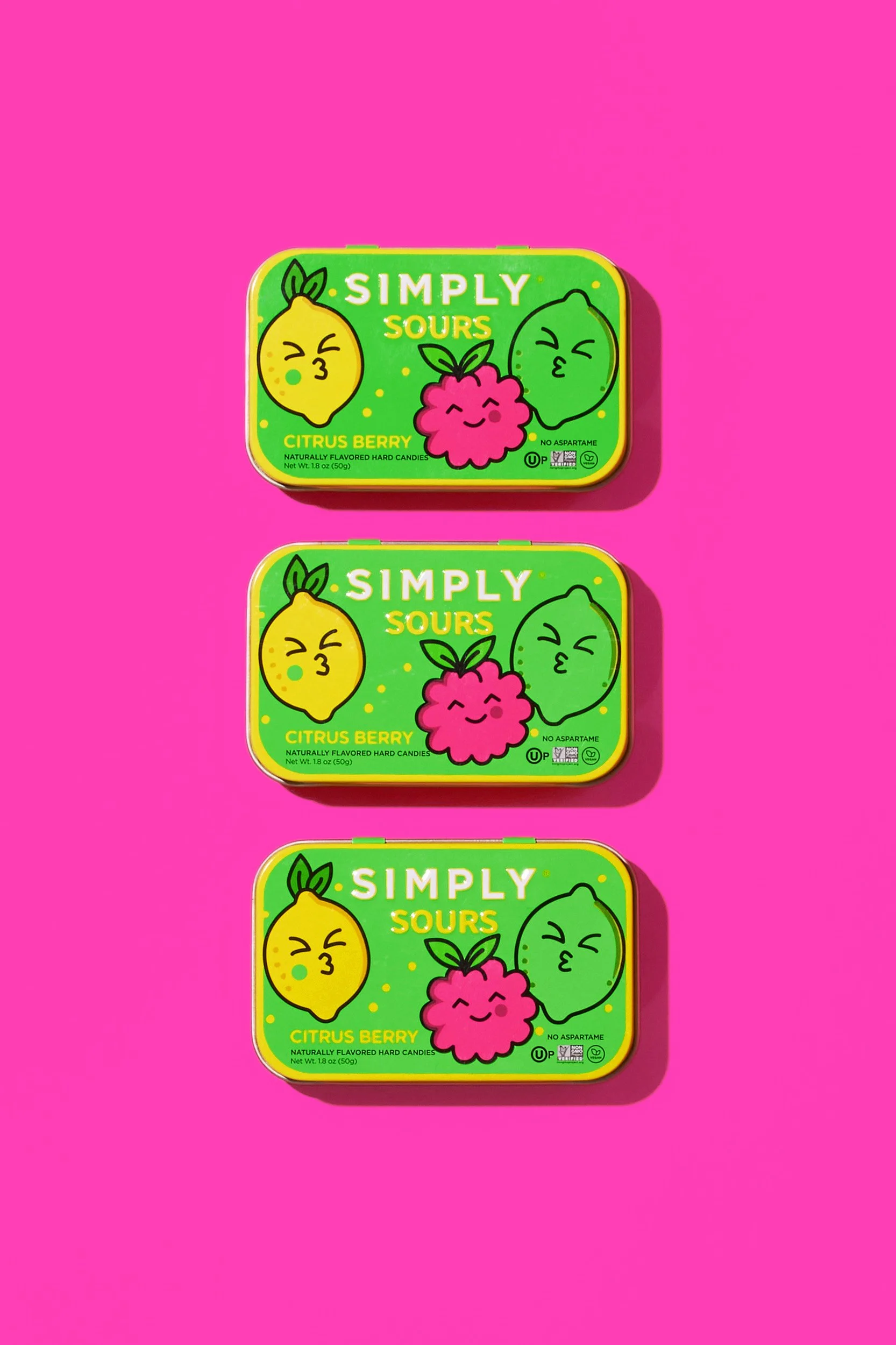 Three rectangular tins of Simply Sours citrus berry flavored hard candies on a pink background, each with cartoon fruit faces.
