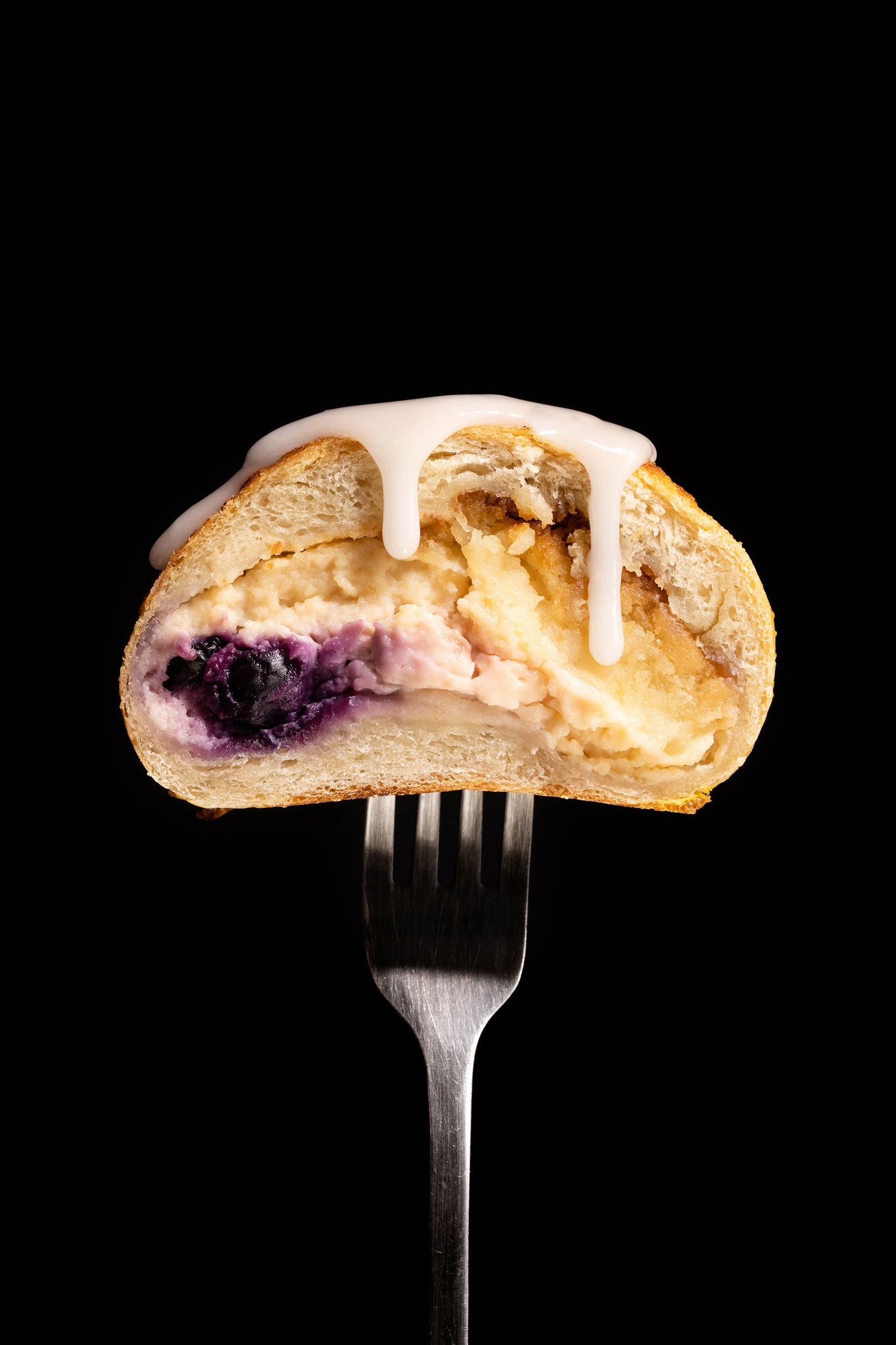 A partially eaten blueberry cheesecake cream-filled pastry with white icing on top, served on a fork against a black background.