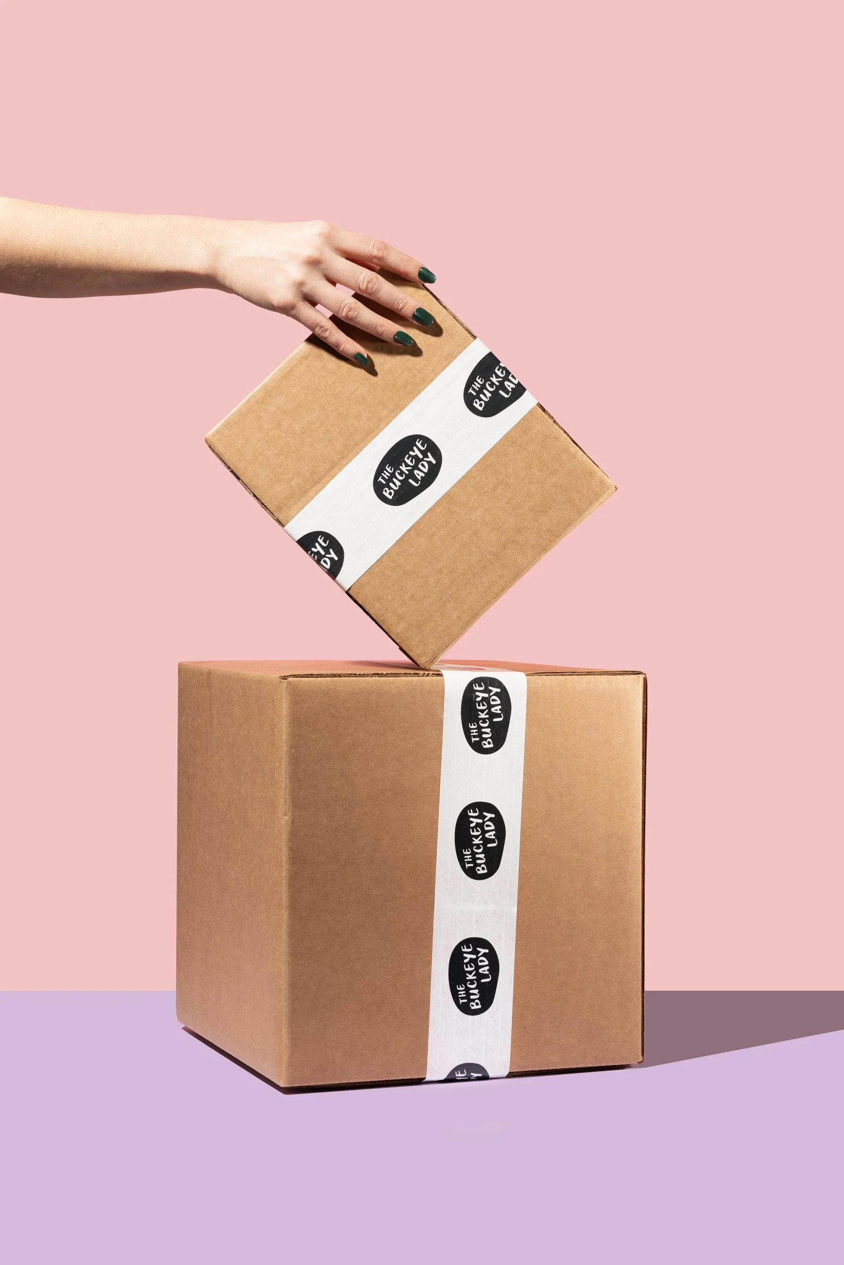 A hand with dark green nails is dropping a smaller cardboard box into a larger cardboard box against a pink and purple background. Both boxes are sealed with white tape that has a black logo reading "The Buckeye Lady."