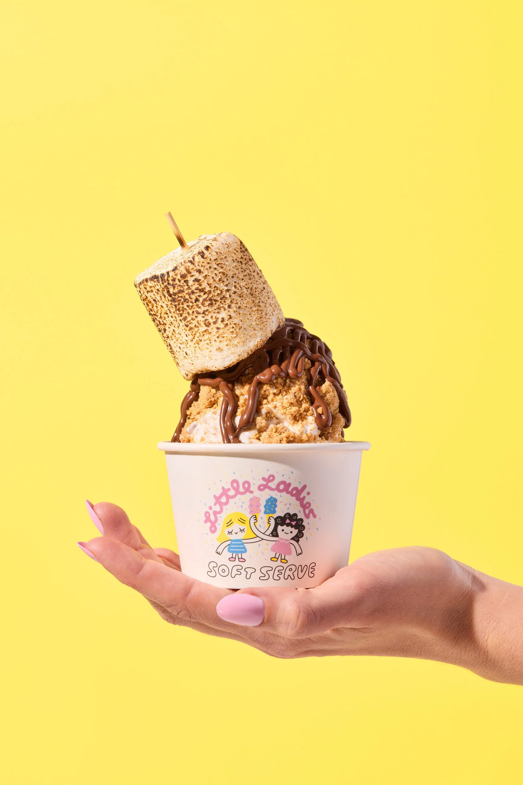 A hand holding a cup of ice cream with a toasted marshmallow, chocolate drizzle, and cookie crumbs against a yellow background.