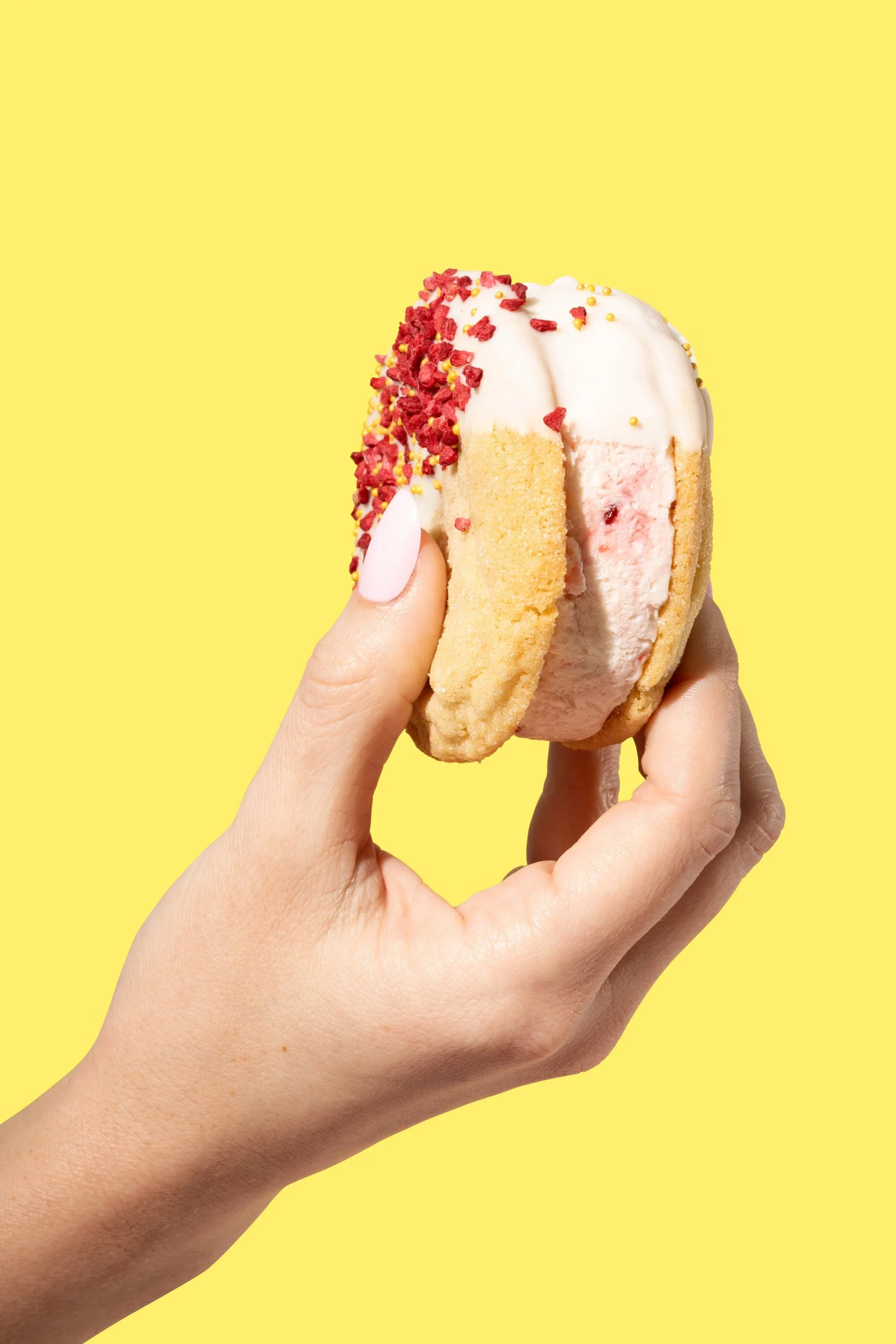A hand holding a half-eaten ice cream sandwich with vanilla ice cream, cookie layers, and red sprinkles against a yellow background.