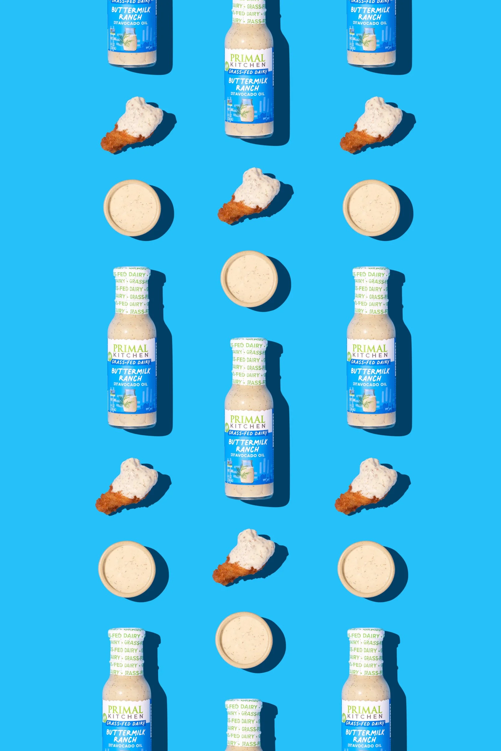 Pattern of bottled Primal Kitchen Buttermilk Ranch dressing, individual sauce cups, and pieces of fried chicken on a bright blue background.